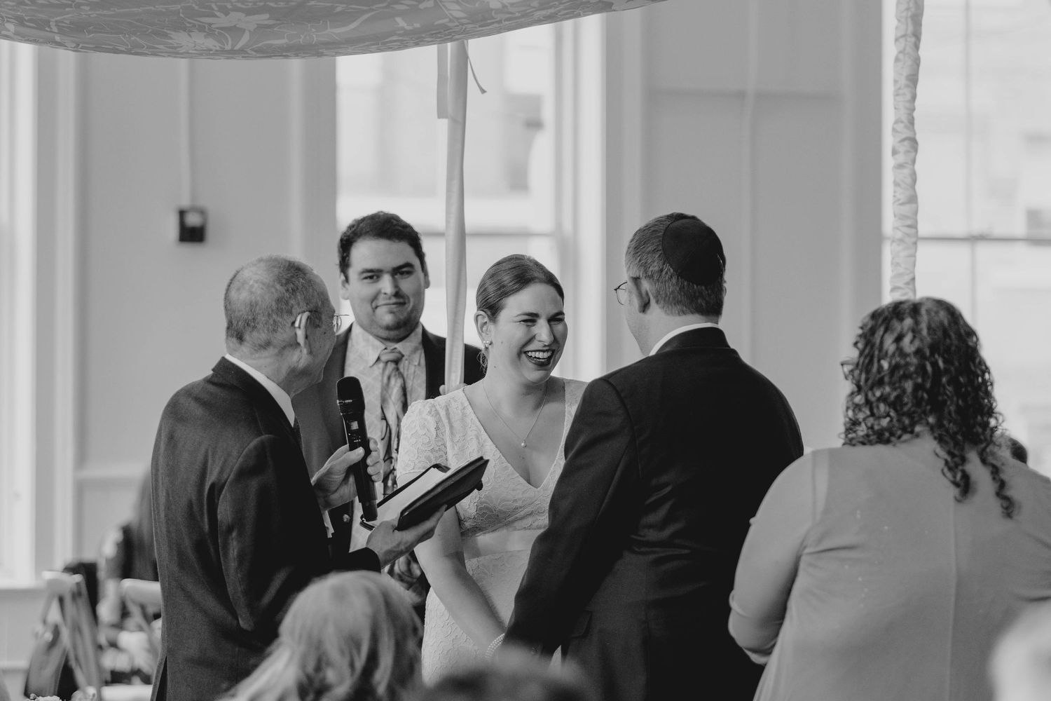 Black and white photo of bride and groom laughing with officiant during intimate indoor wedding ceremony.