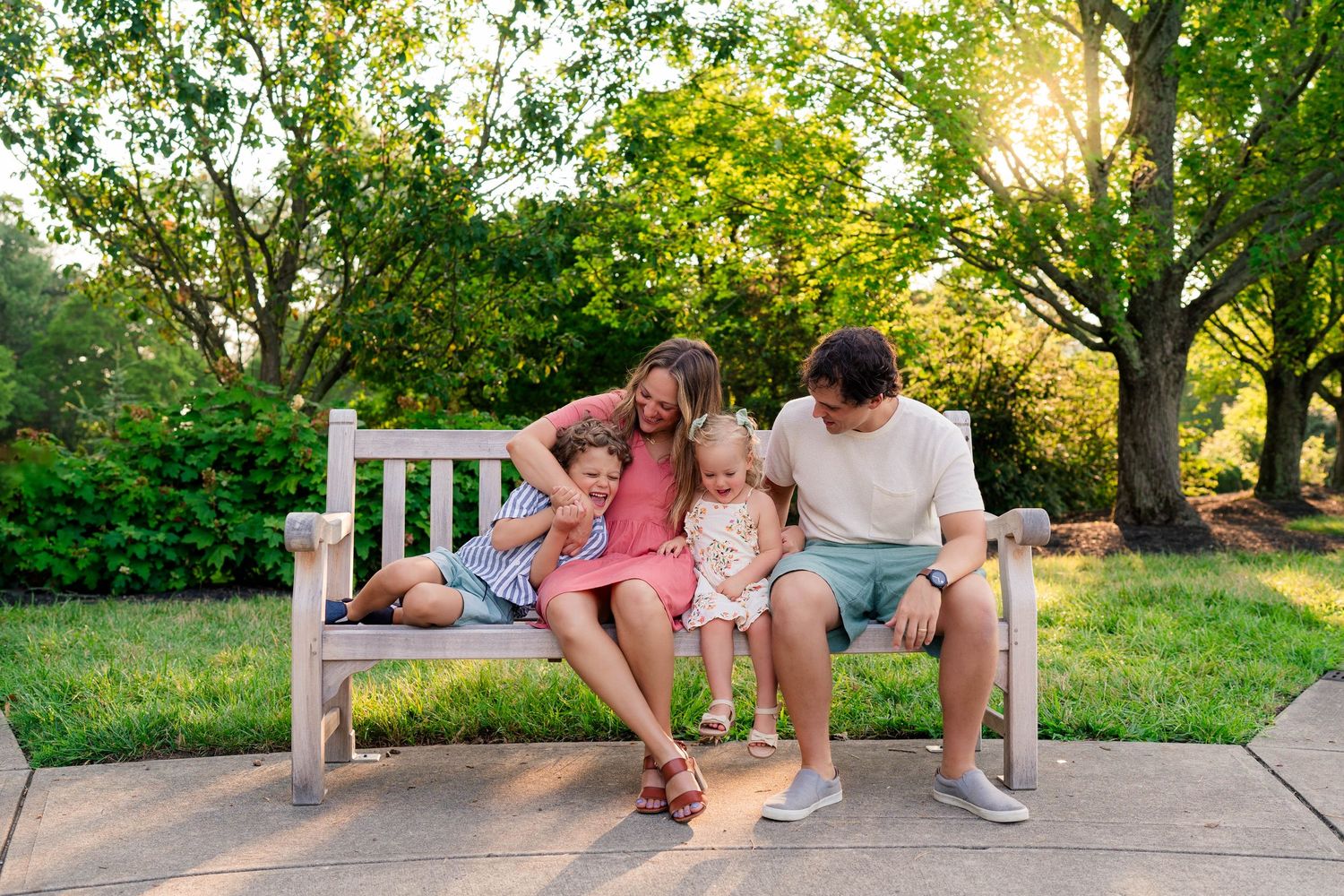 Family of four sitting on a white park bench surrounded by lush green trees in golden hour sunlight.