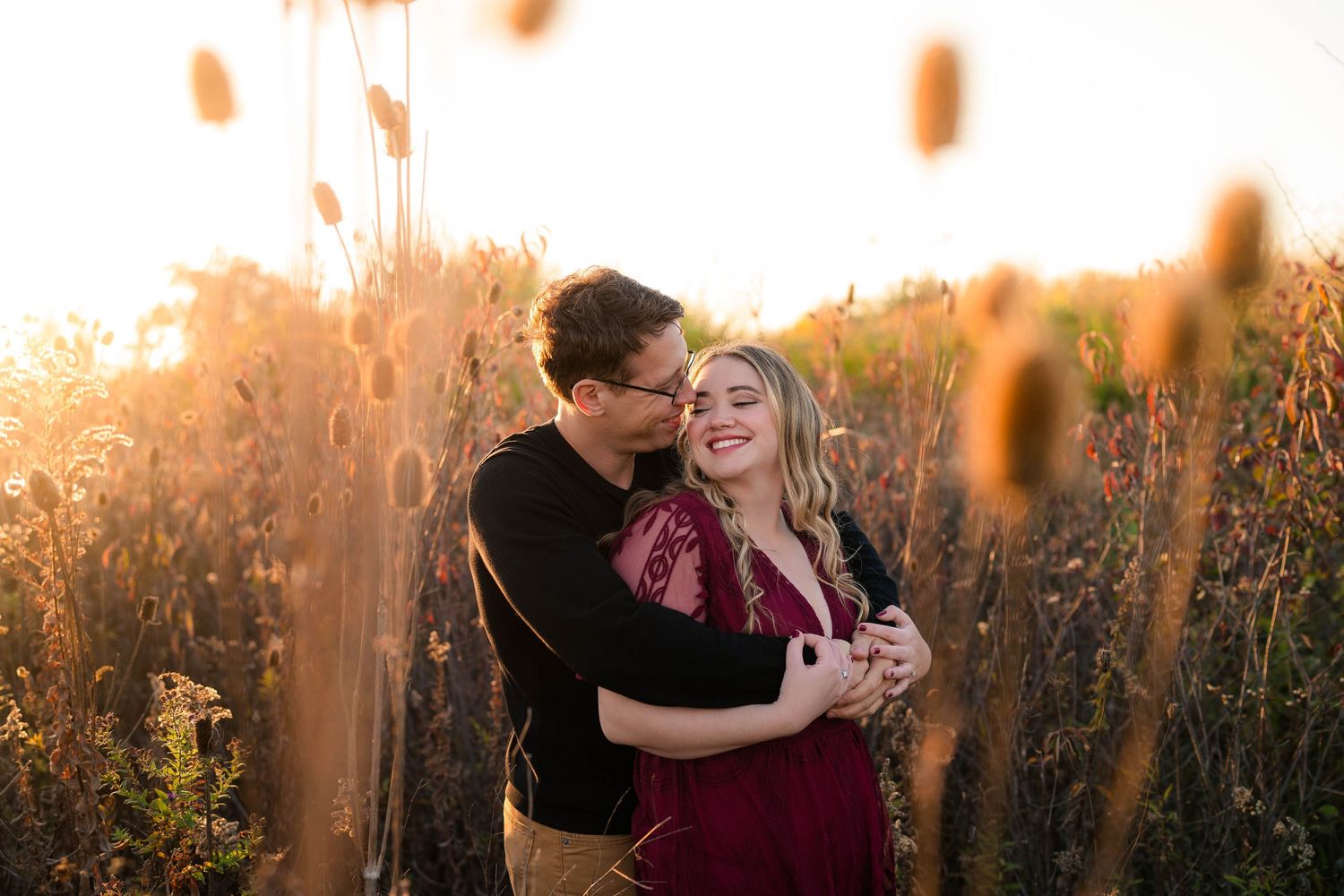 Couple embracing in golden field at sunset, man kissing woman's cheek as she smiles joyfully.
