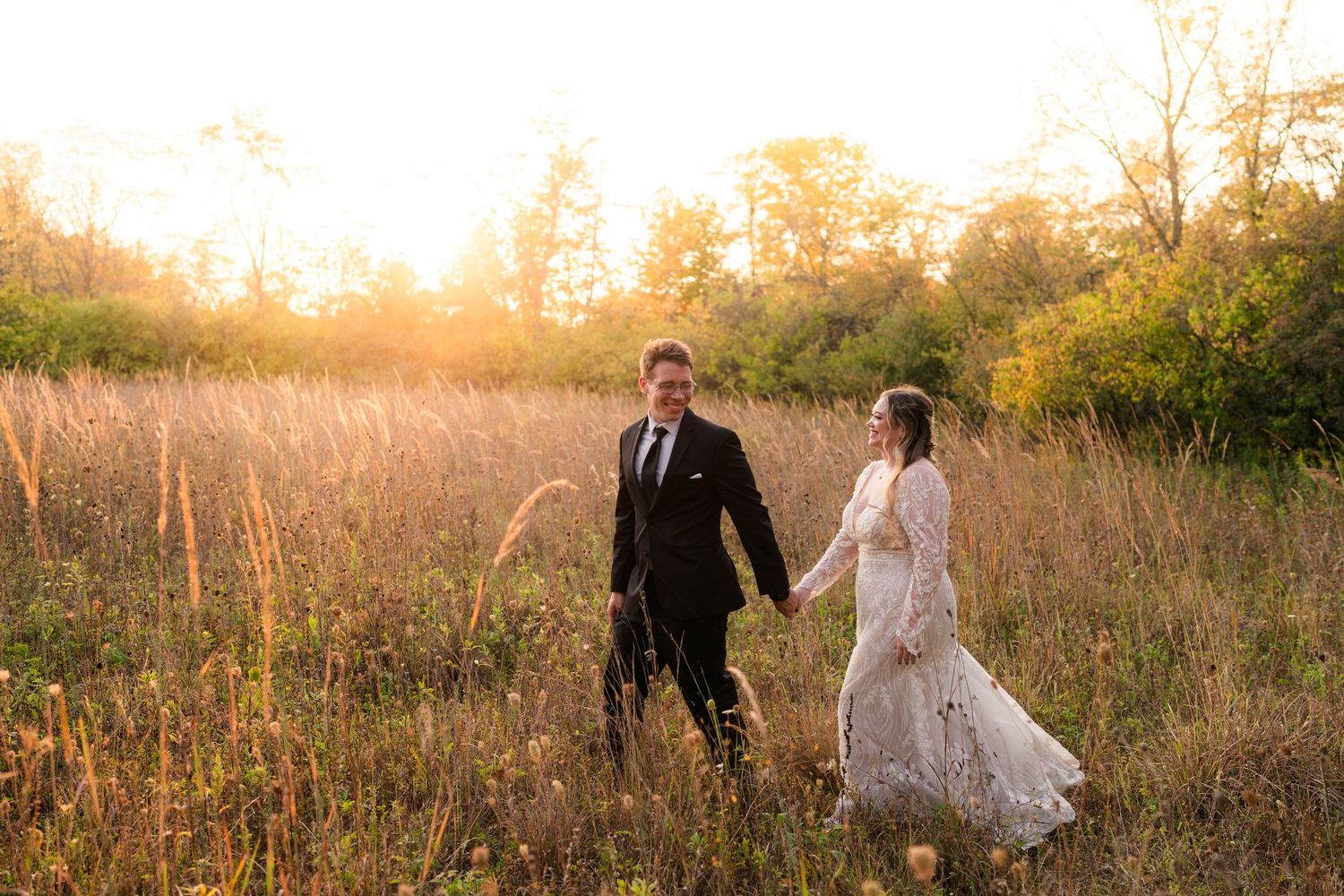 Newlyweds share a romantic moment walking through a sunlit meadow in wedding attire.