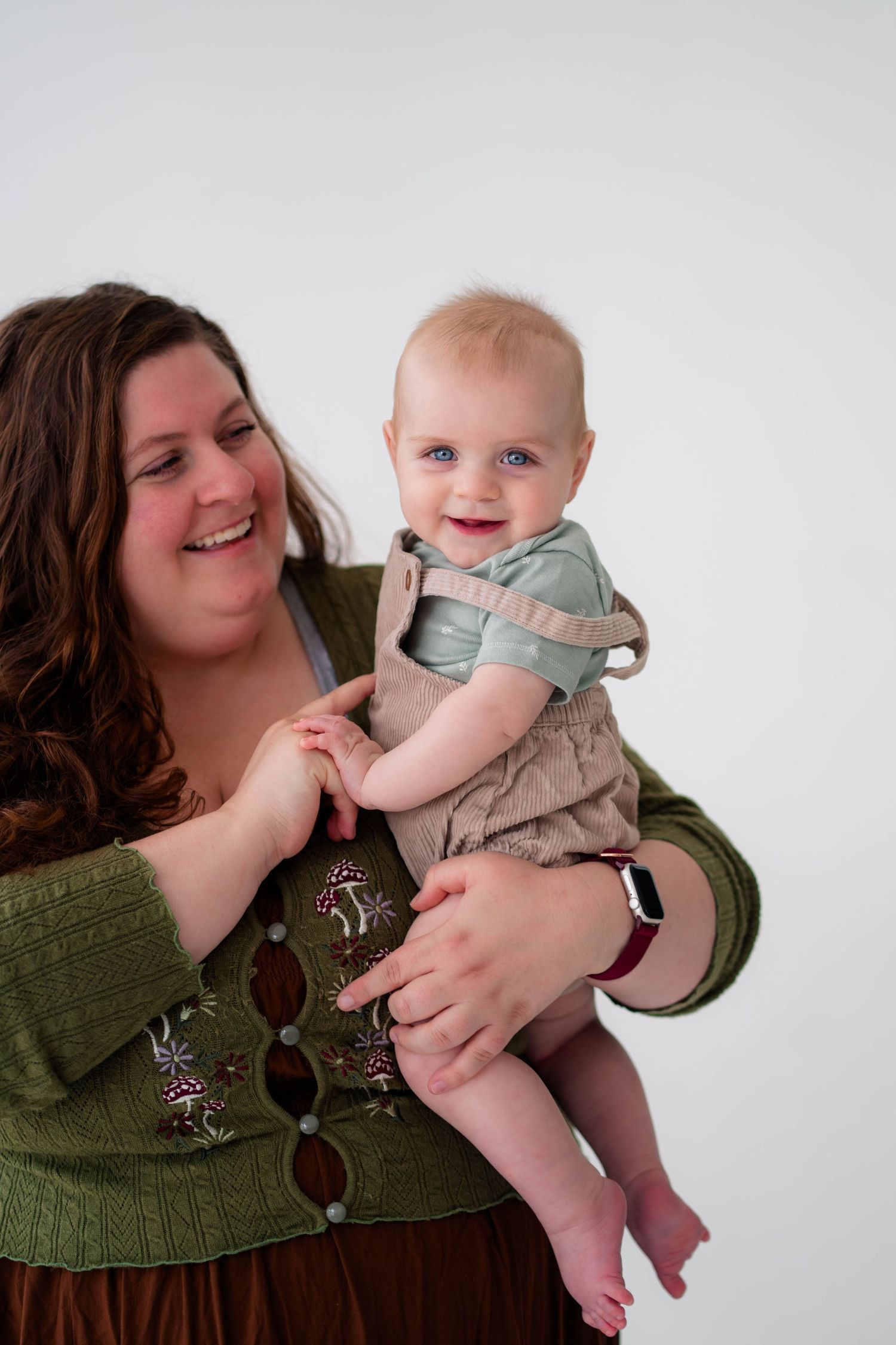 Joyful mother in green top holding laughing blue-eyed baby in tan overalls on white background.