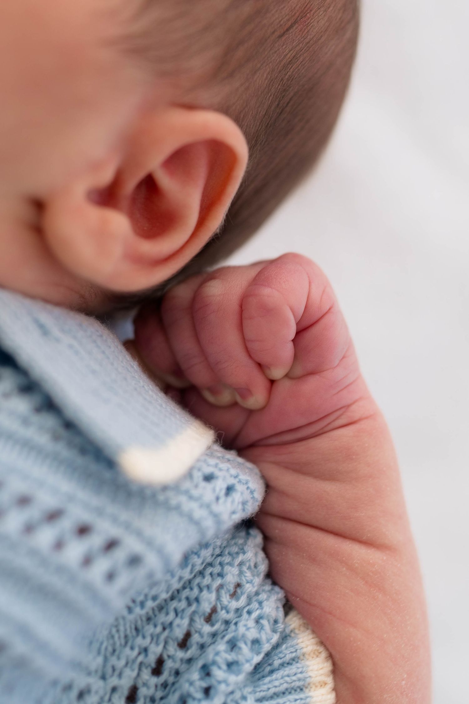 A close-up detail shot of a newborn babys ear and tiny clenched fist resting against a soft blue knit outfit.