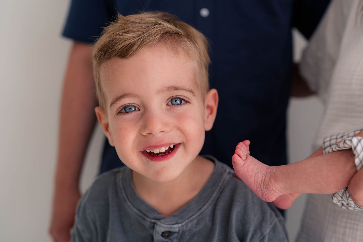 Smiling toddler boy with blue eyes and blonde hair in a gray shirt during a family photo session.