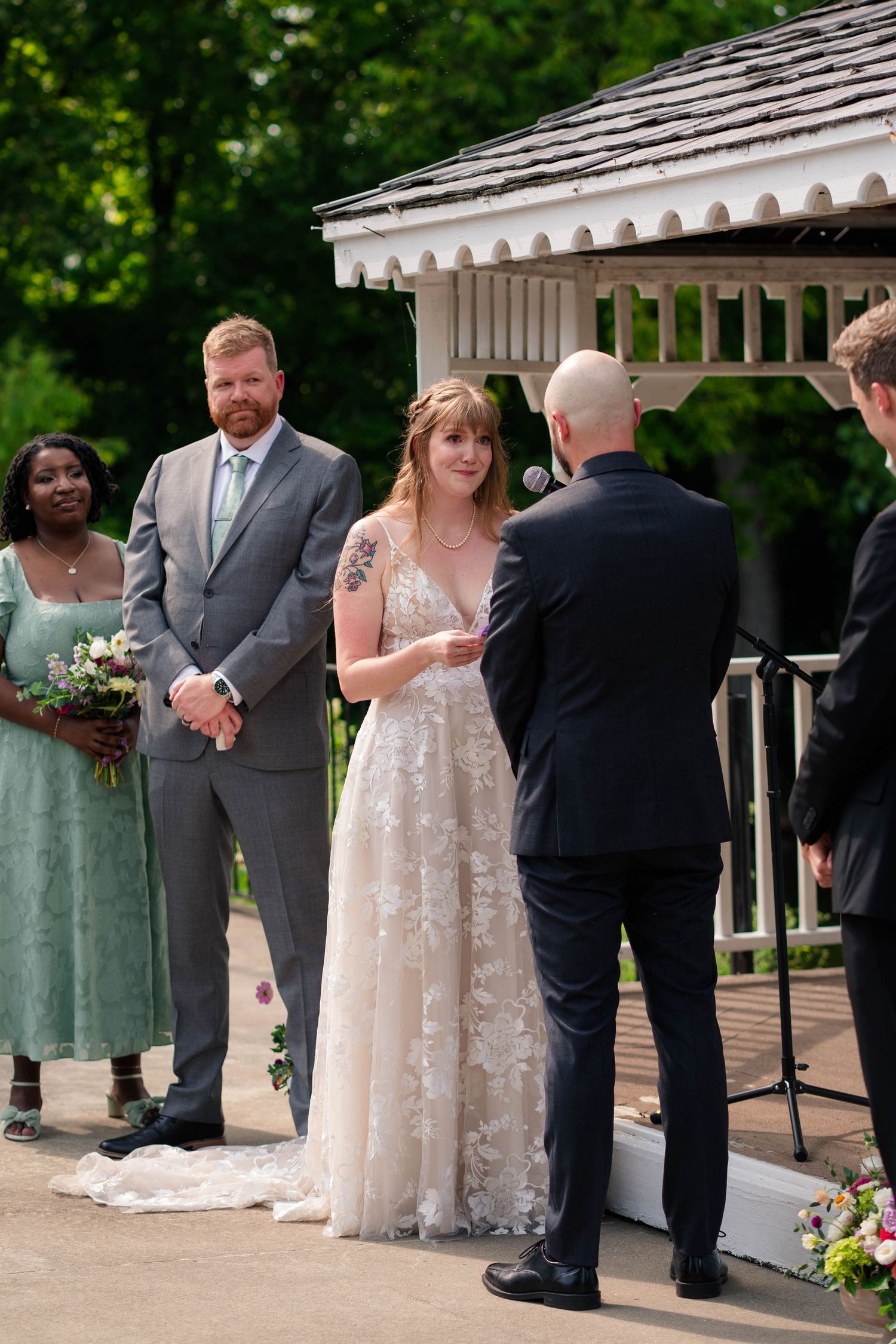Bride in lace gown reads vows to groom at outdoor gazebo wedding ceremony with wedding party standing nearby.