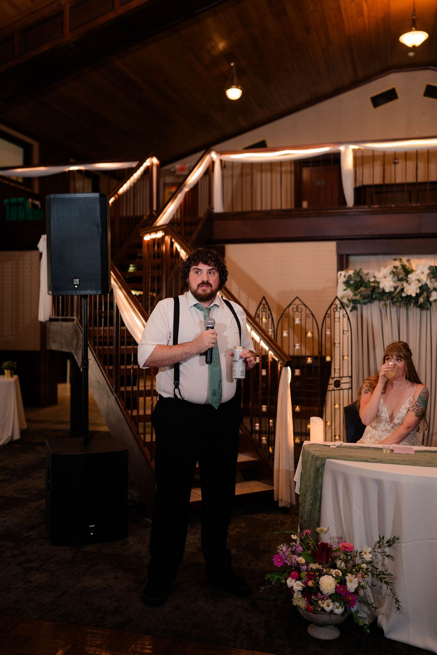 Groomsman in suspenders giving a toast with microphone at an elegant wedding reception.