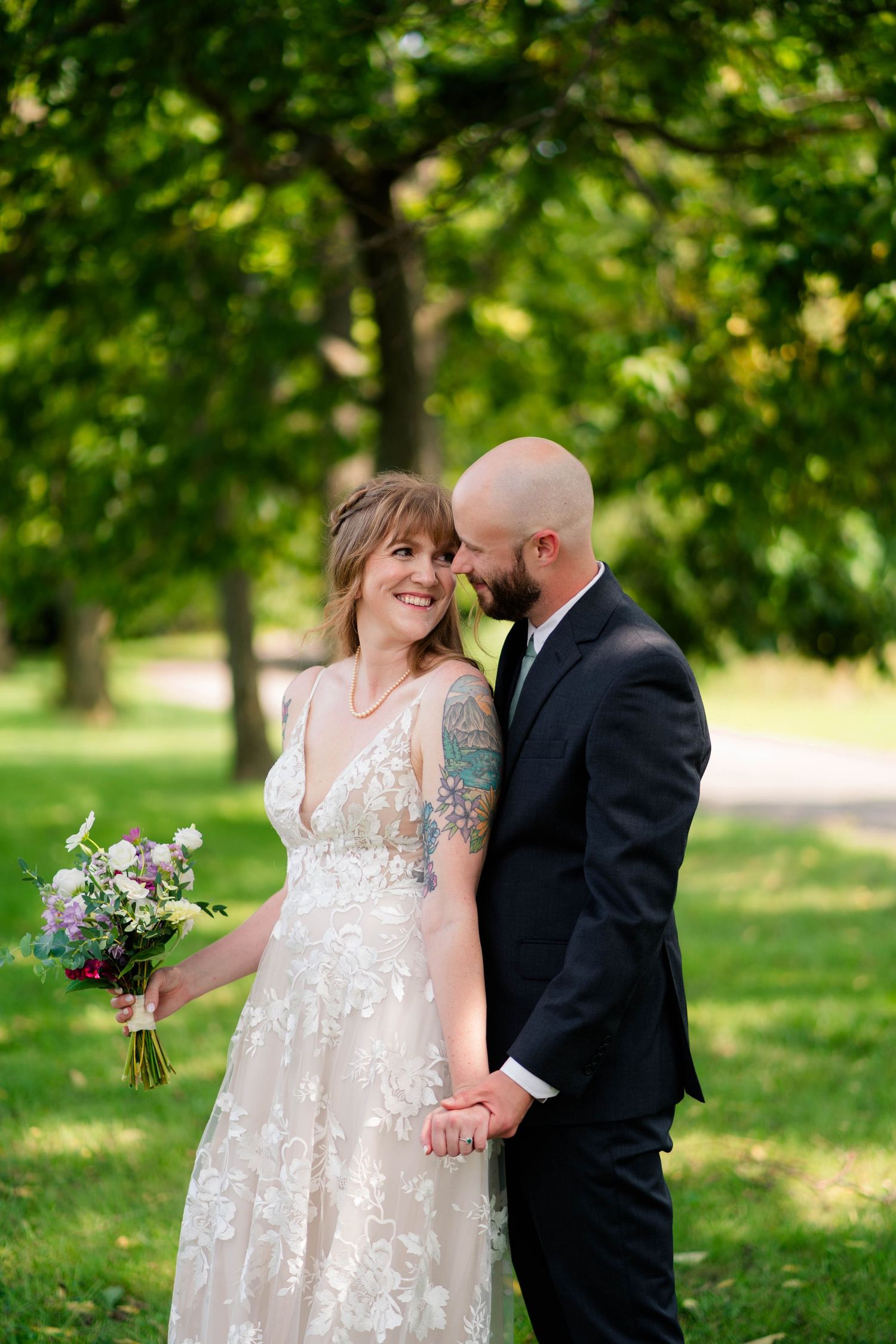 A bride with a tattooed arm and groom hold hands and smile together in a lush green outdoor setting.