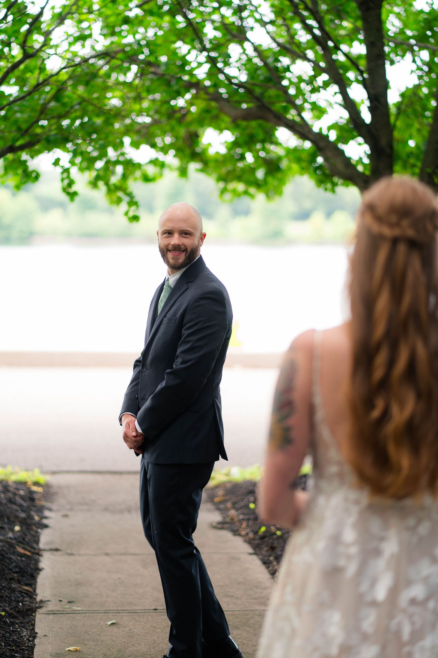 Groom in dark suit smiling at bride during outdoor first look by lakeside.