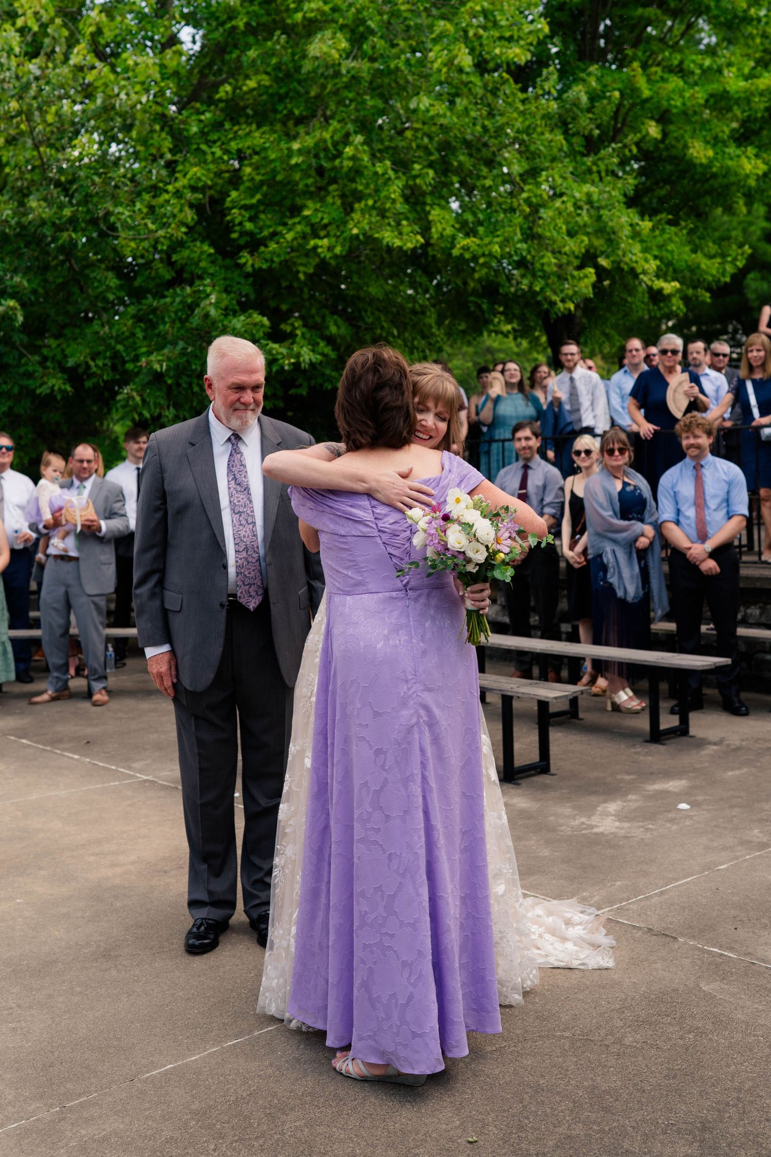 Bride hugging woman in purple gown during outdoor wedding ceremony with guests watching.