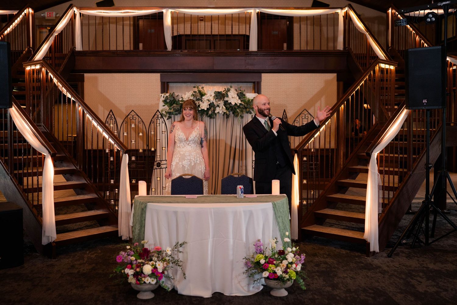 Bride and bald groom stand at sweetheart table beneath grand staircase at elegant wedding reception.