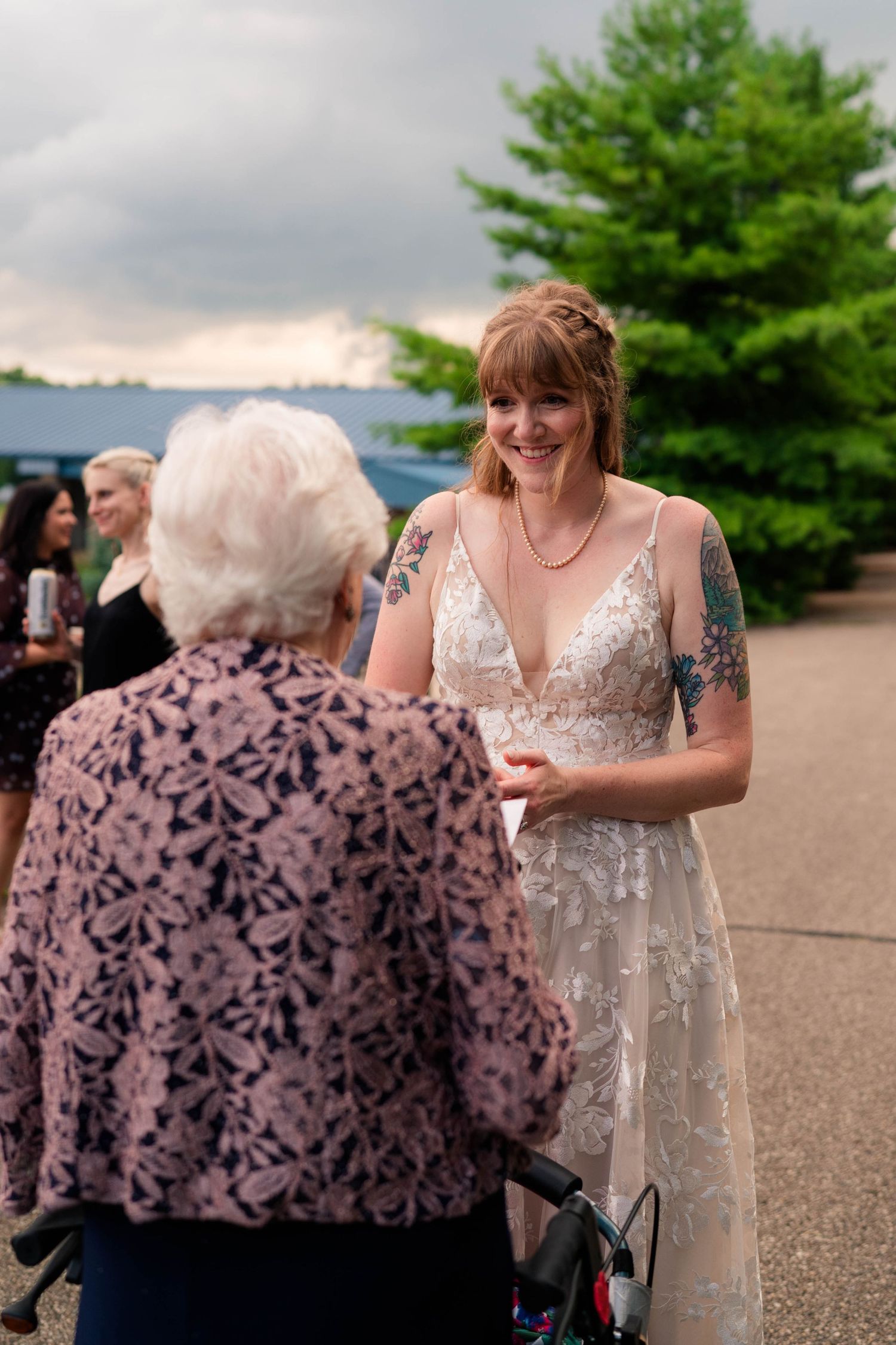Smiling bride in a lace wedding gown joyfully greets an elderly woman with a walker outdoors.