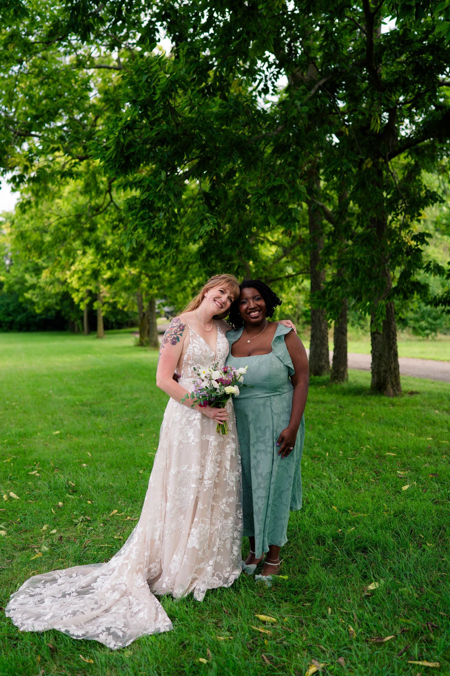 Bride in lace gown holds bouquet while posing with bridesmaid in sage green dress outdoors.