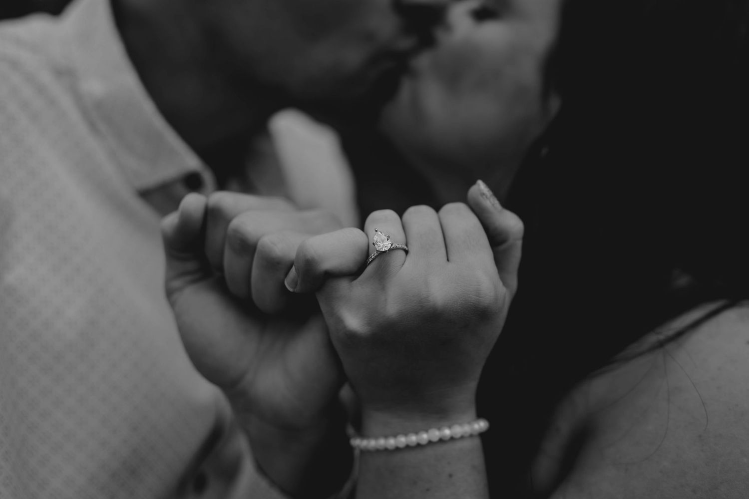 Black and white close-up of engaged couple kissing while holding hands showing diamond ring.