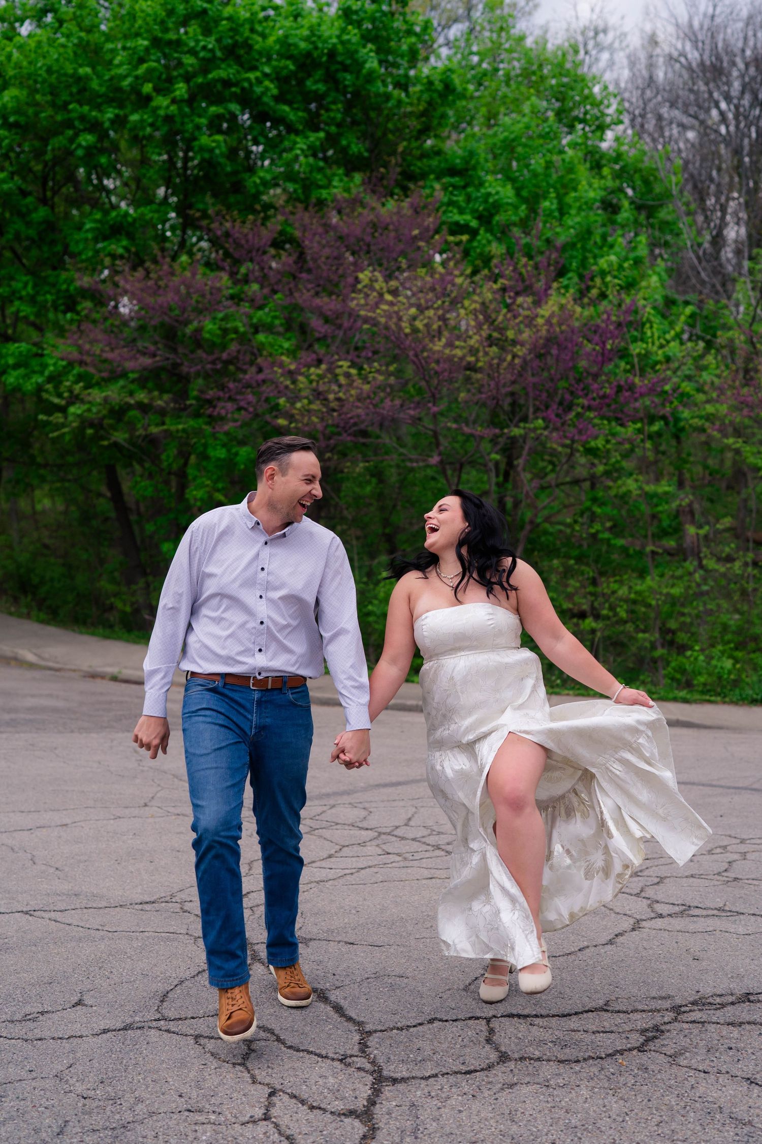 Joyful couple laughing and holding hands while walking on pavement with colorful trees behind.
