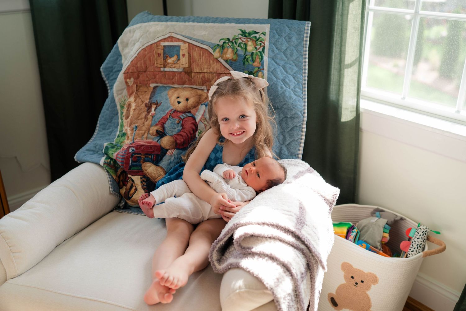 Young girl sits in armchair holding newborn baby, with teddy bear quilt backdrop and toys nearby.