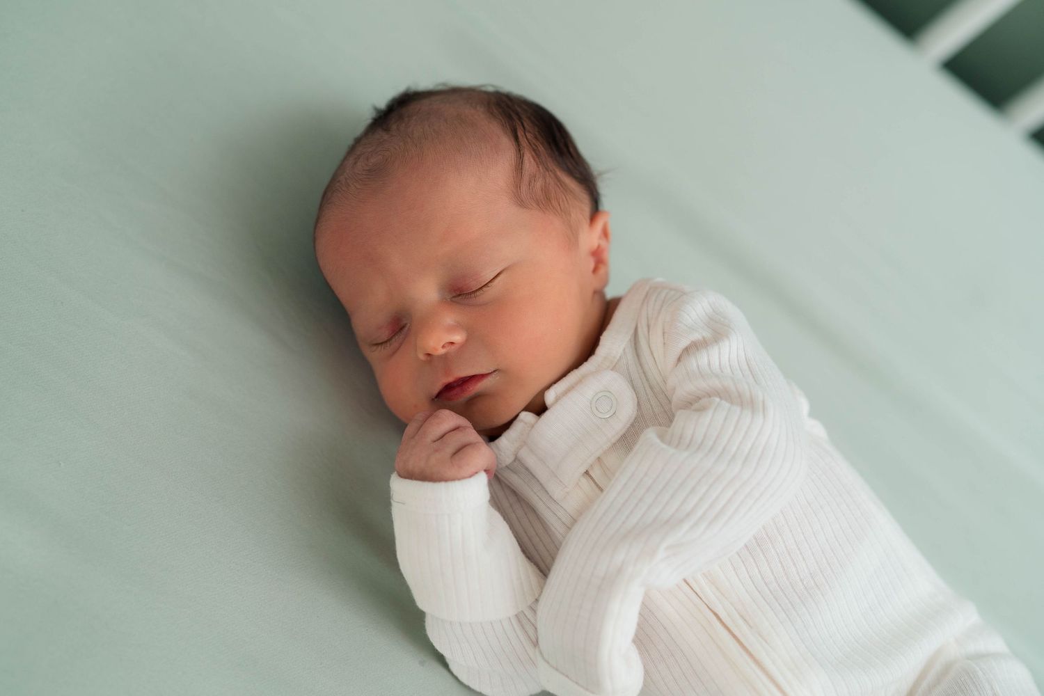 A peaceful newborn baby sleeps soundly in a white ribbed onesie on a mint green surface.