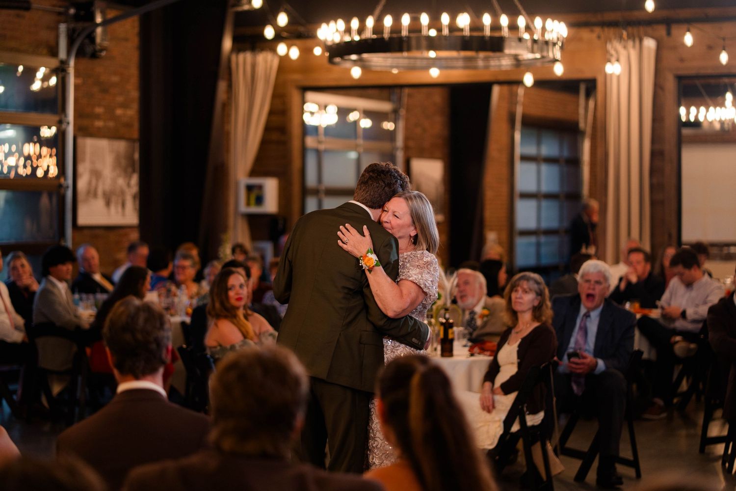 Bride and groom share intimate first dance surrounded by seated guests in warmly lit wedding venue.