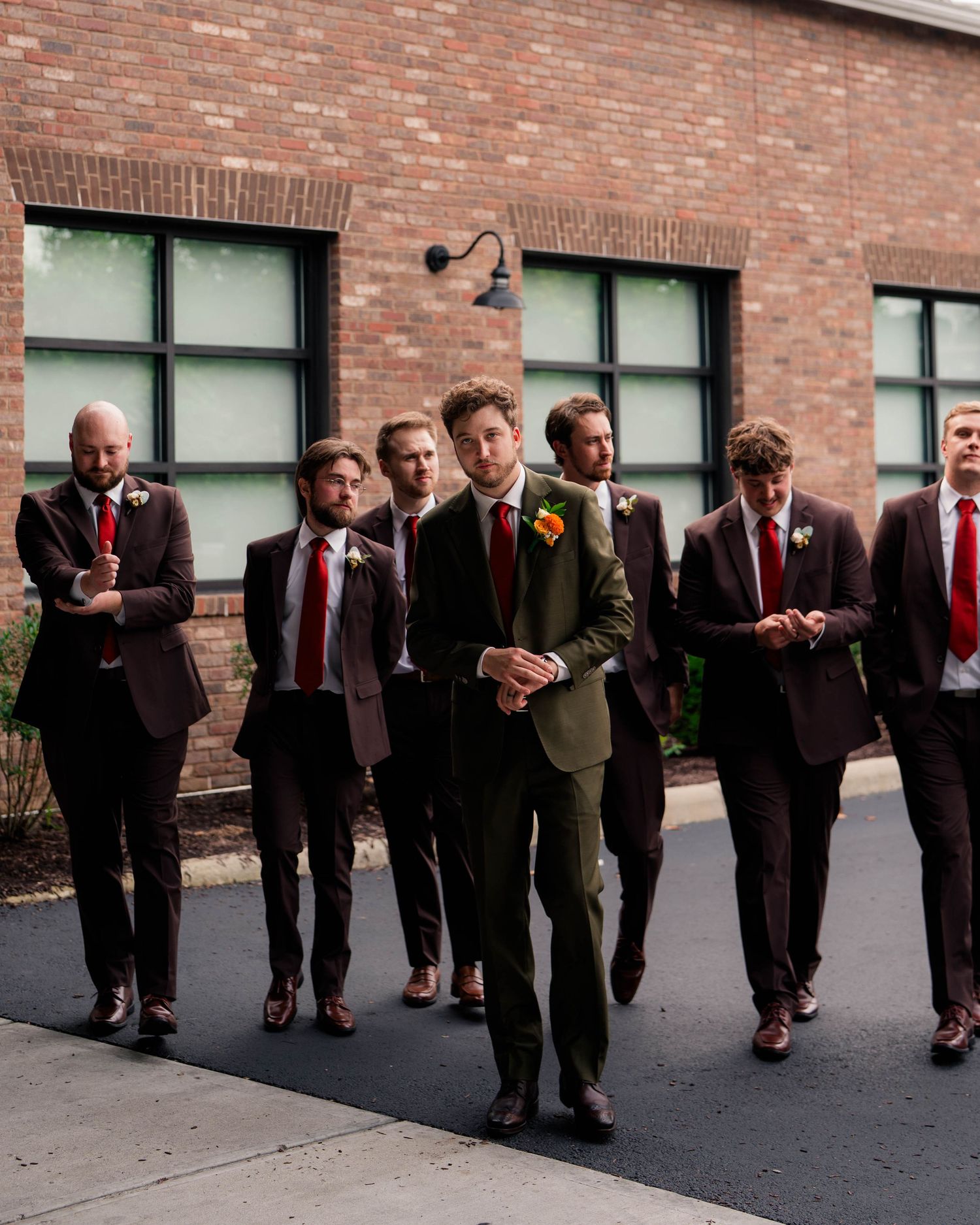 Groom in olive suit leads groomsmen in burgundy suits walking outside brick building.