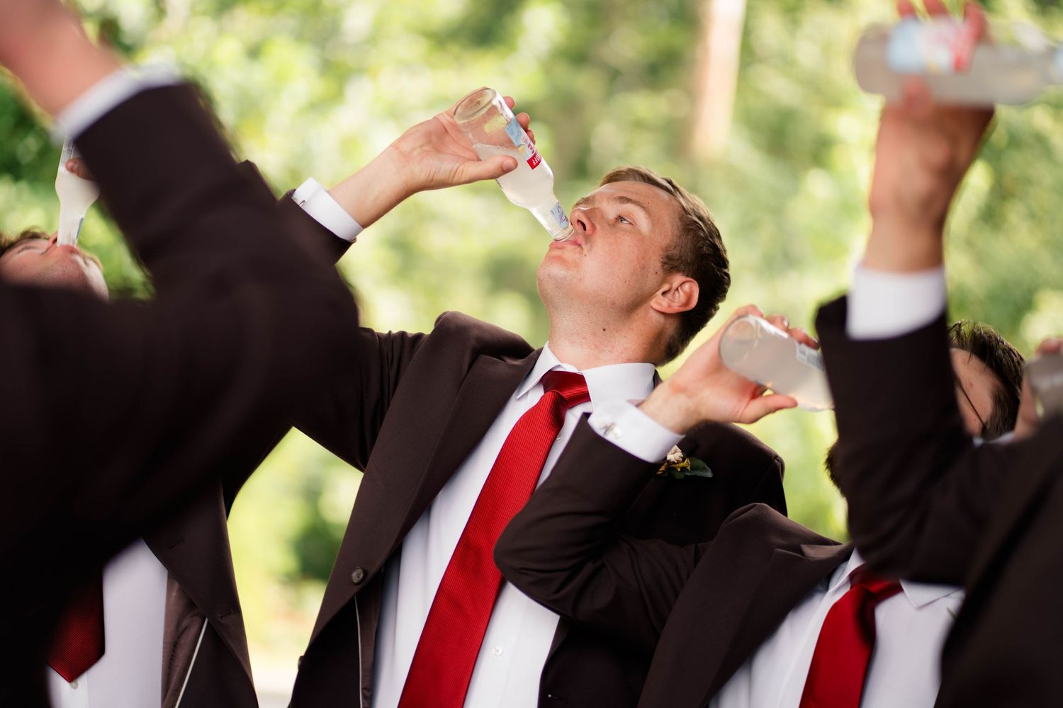 Groomsmen in dark suits with red ties doing shots outdoors at wedding celebration.