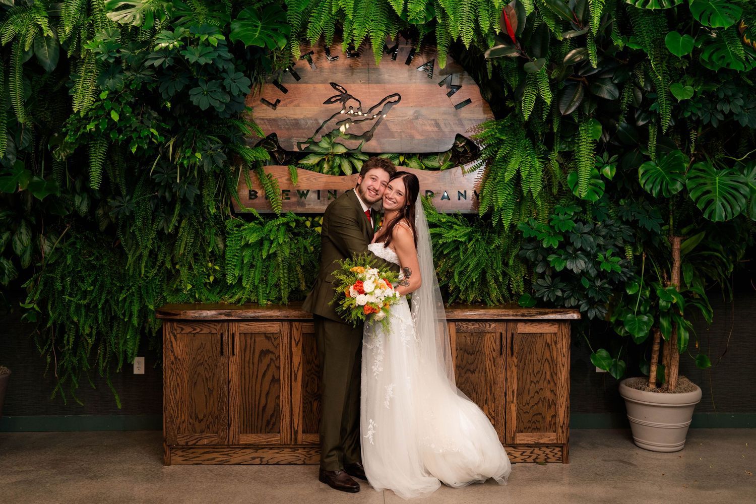 Bride and groom embrace in front of lush green botanical wall at elegant indoor wedding venue.