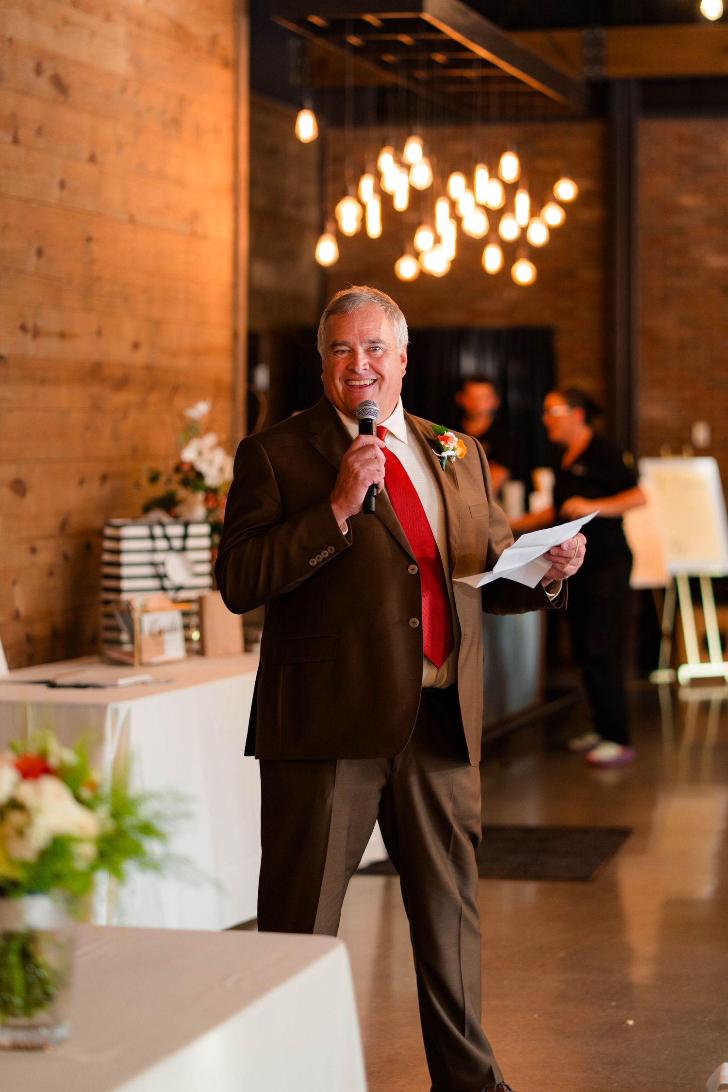Smiling man in brown suit and red tie delivering wedding toast speech with microphone.