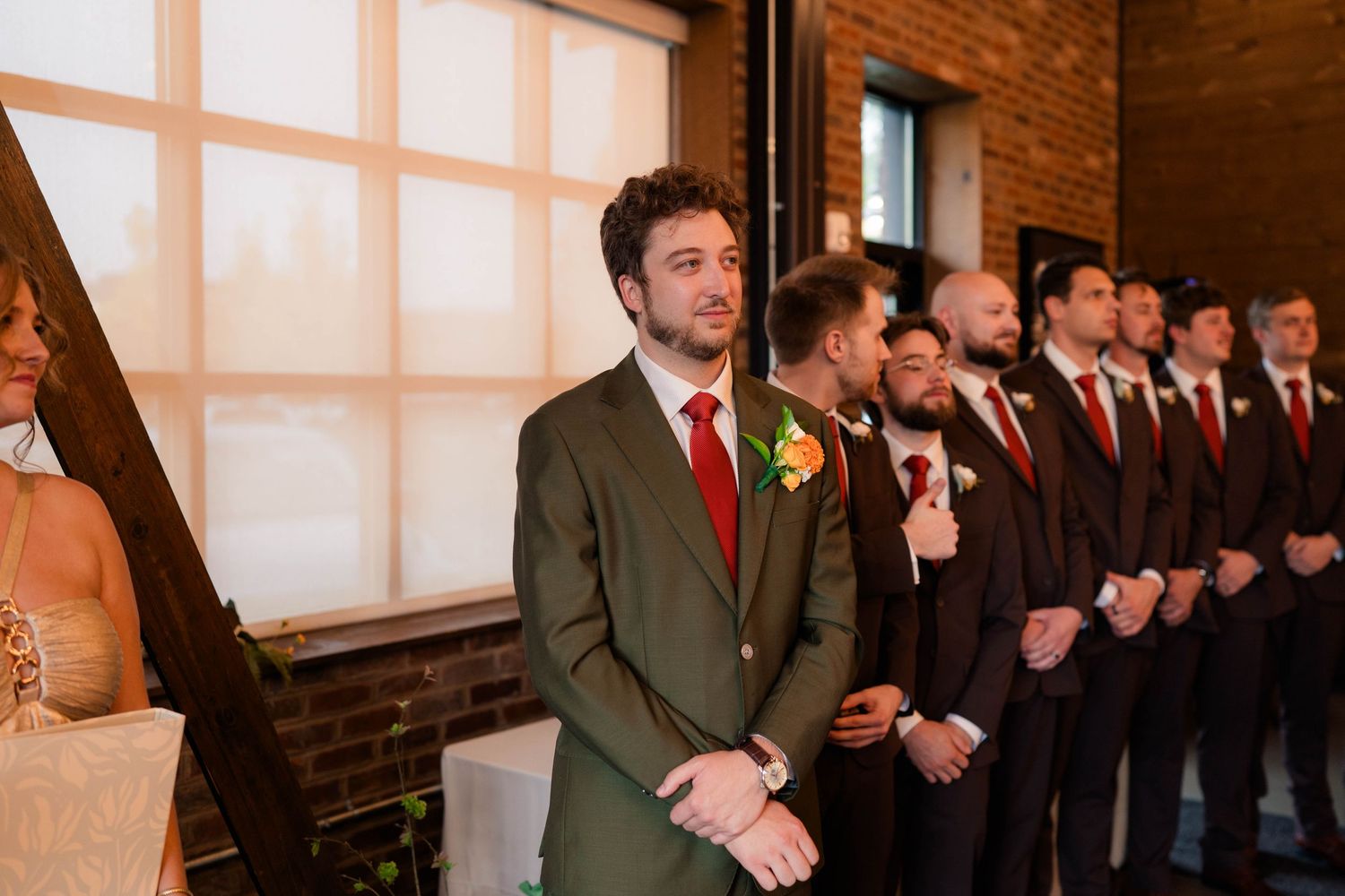 Emotional groom in olive suit with red tie watches bride approach at indoor wedding ceremony.