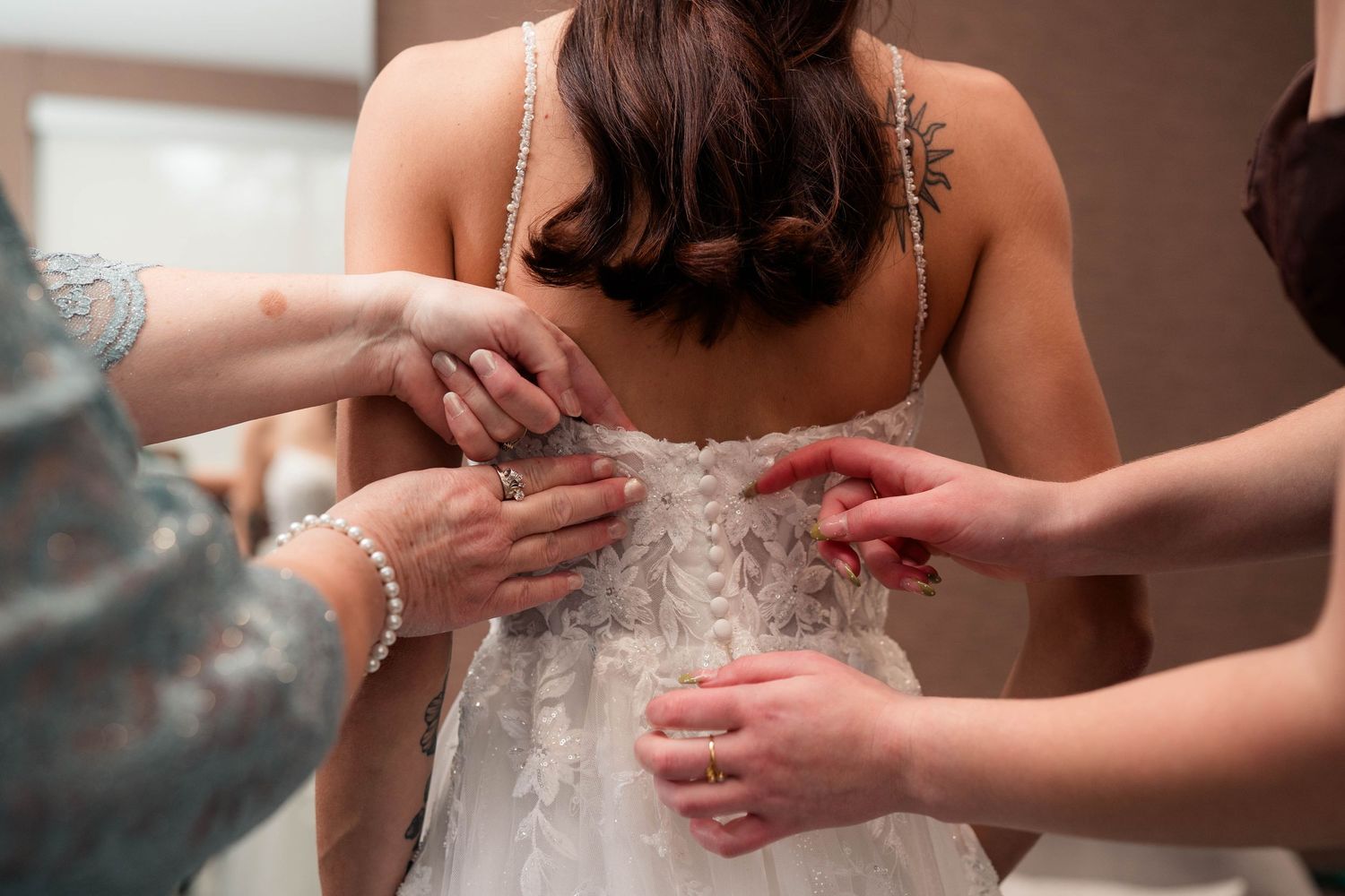 Bridesmaids helping bride fasten lace wedding dress from behind in bridal preparation room.