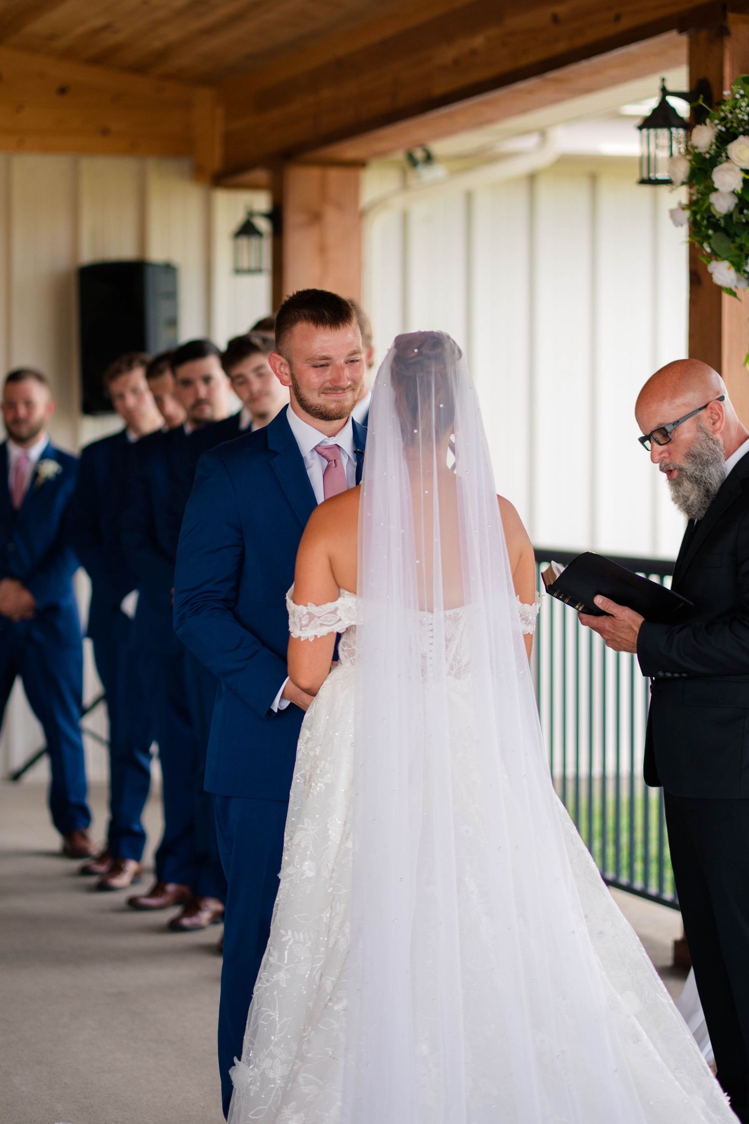 Groom in navy suit smiles at bride during wedding vows ceremony with groomsmen lined up behind.