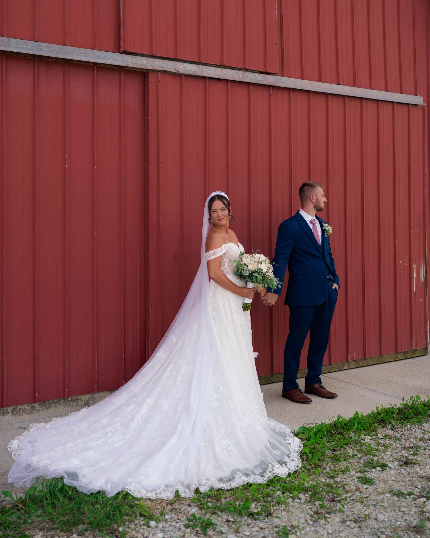 Bride in off-shoulder lace gown with cathedral train stands beside groom in navy suit by red barn.