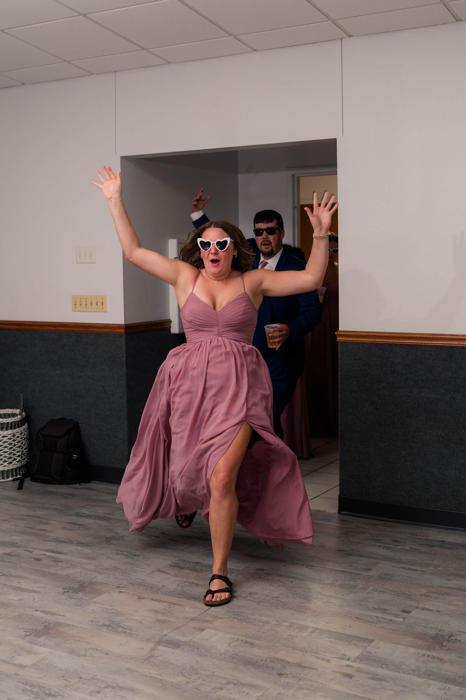 Excited bridesmaid in pink dress and heart sunglasses makes grand entrance at wedding reception.