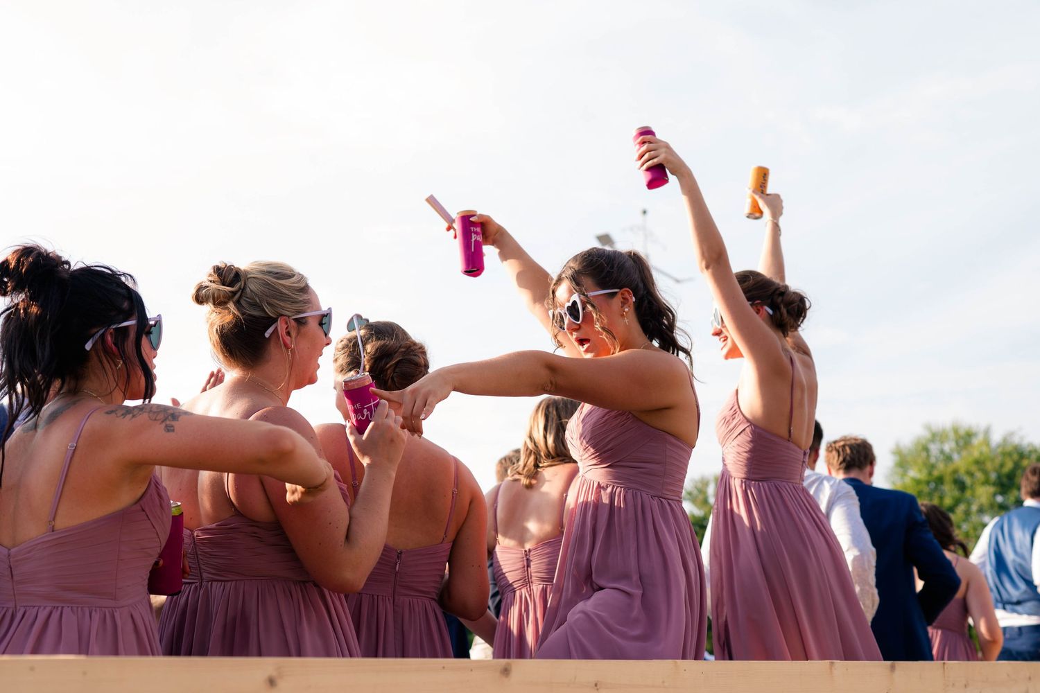 Bridesmaids in mauve dresses raise drinks and celebrate together at outdoor wedding reception party.