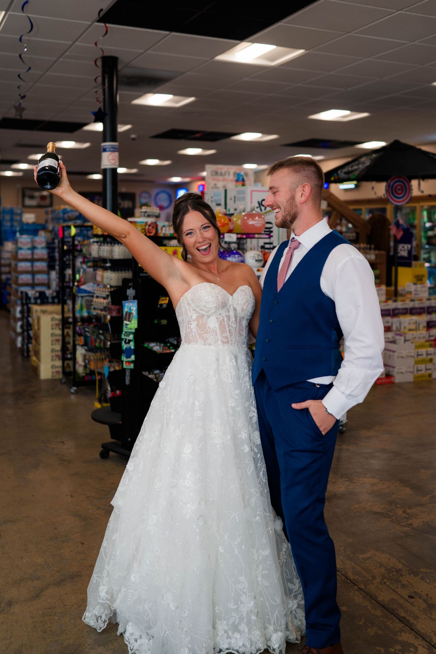 Laughing bride in lace gown raises bottle while groom in navy vest smiles inside liquor store.