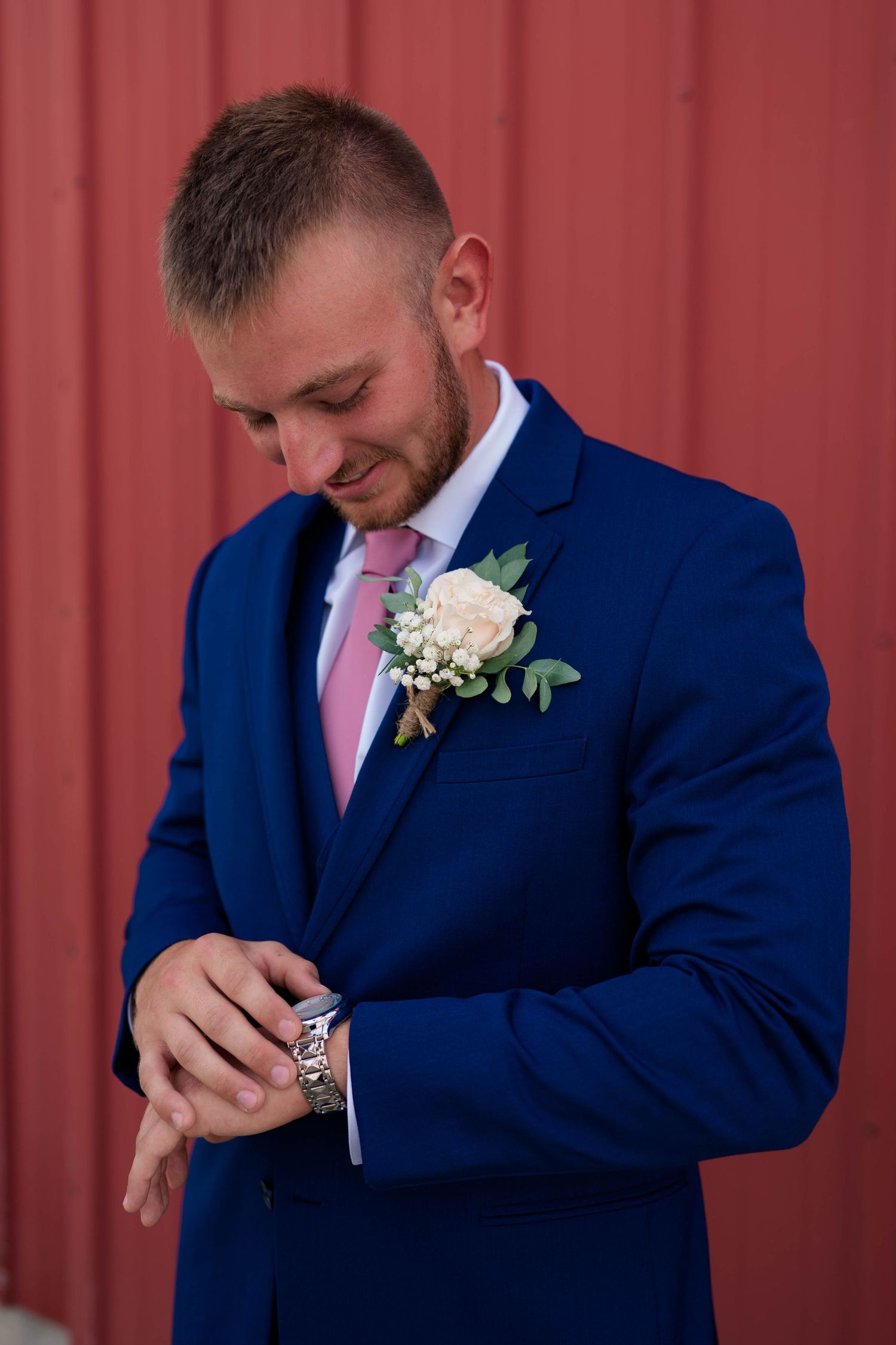 Groom in navy blue suit with pink tie and boutonniere checking his watch before wedding ceremony.