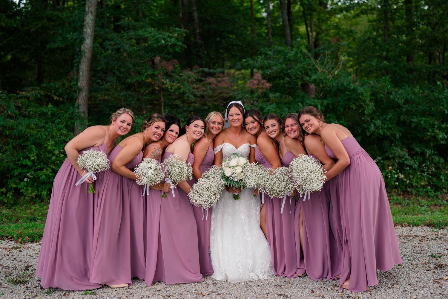 Bride surrounded by bridesmaids in mauve gowns holding baby's breath bouquets outdoors.