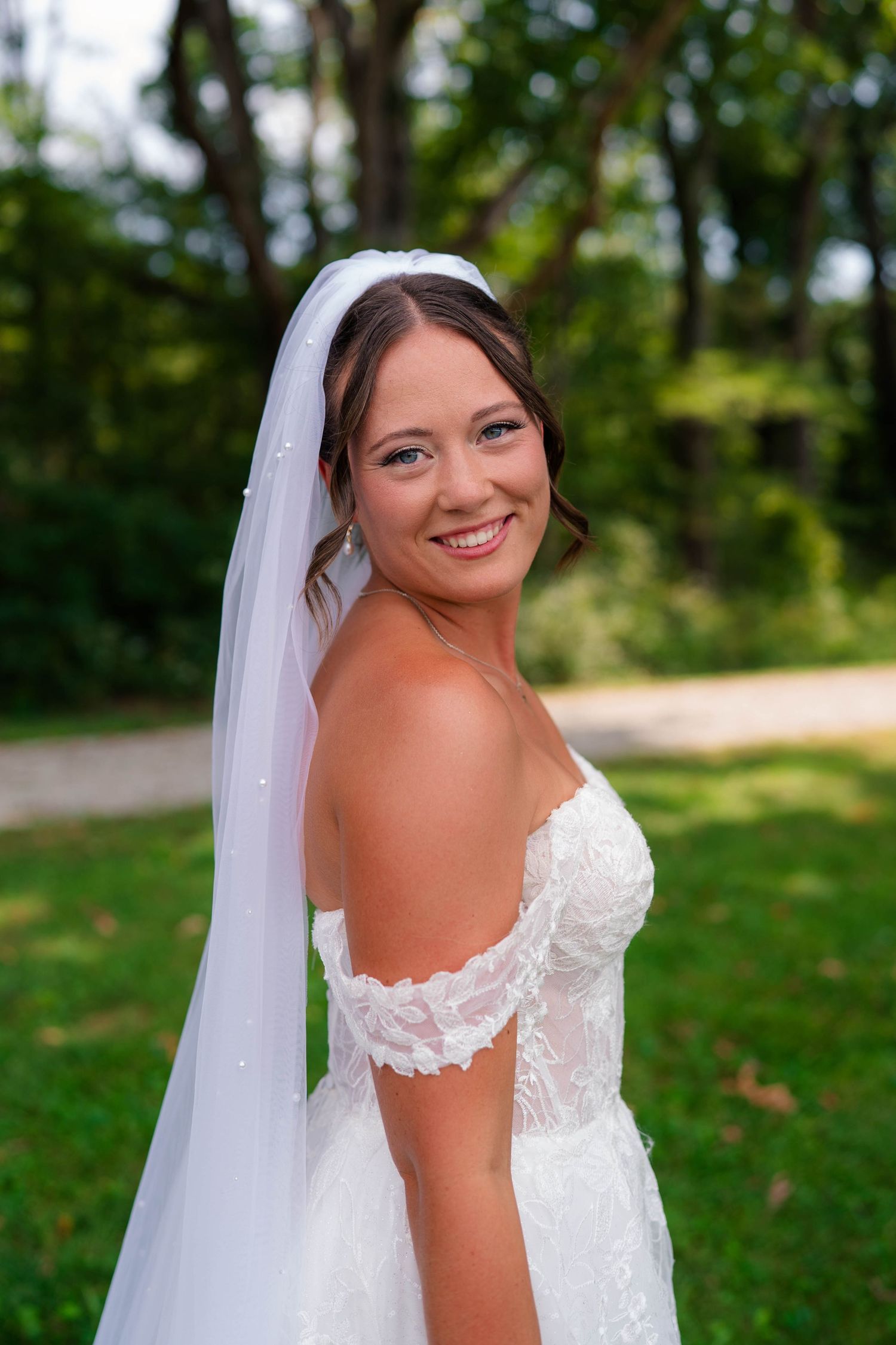 Beautiful bride in off-shoulder lace wedding gown and veil smiles outdoors on her wedding day.