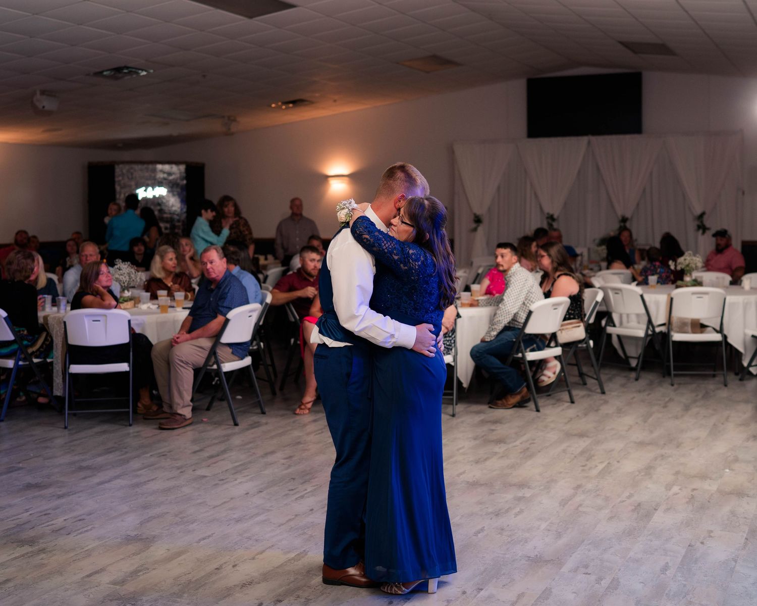 Couple shares romantic first dance in blue formal wear at wedding reception with guests seated at round tables.