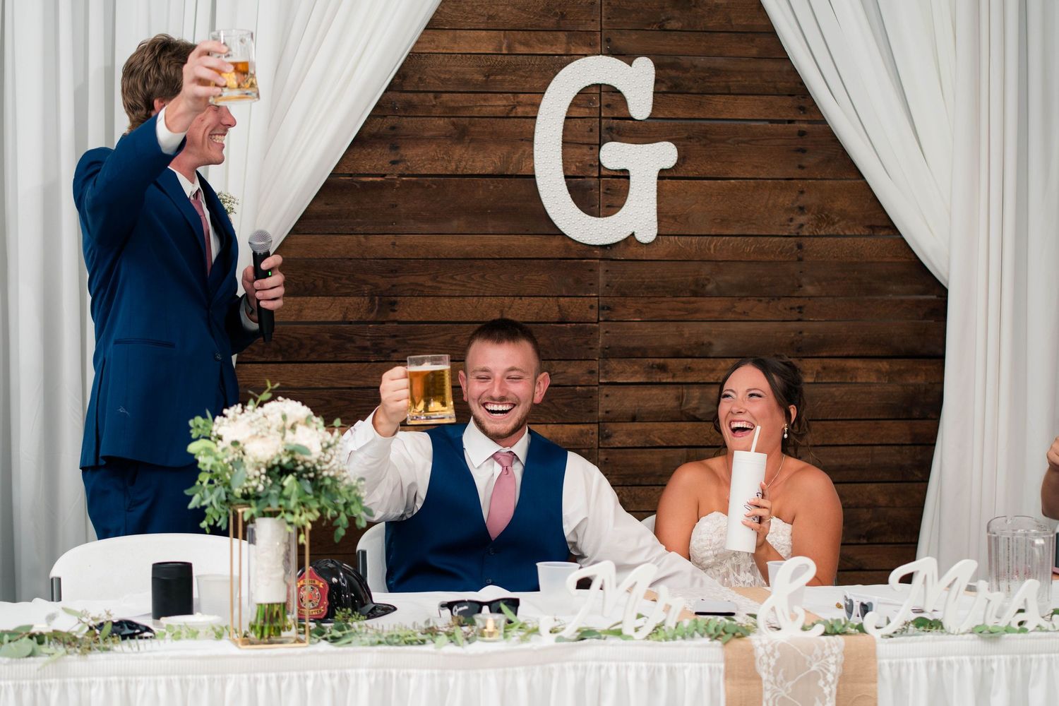 Groom and bride laugh as they raise glasses for a toast at their wedding reception.