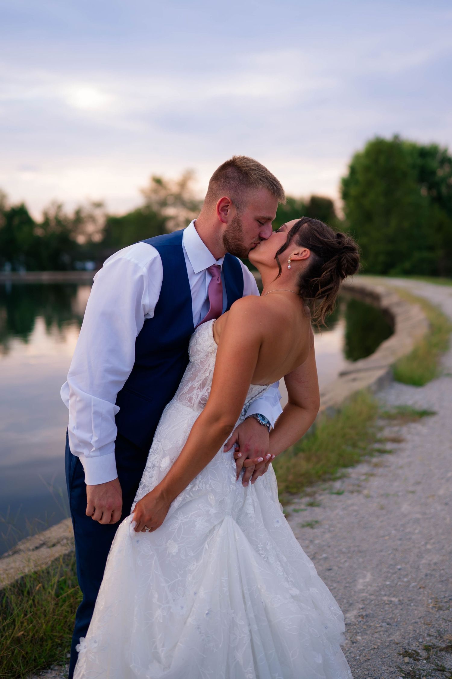 Bride and groom share a passionate kiss by a lakeside path at sunset, groom in navy vest and bride in white lace gown.