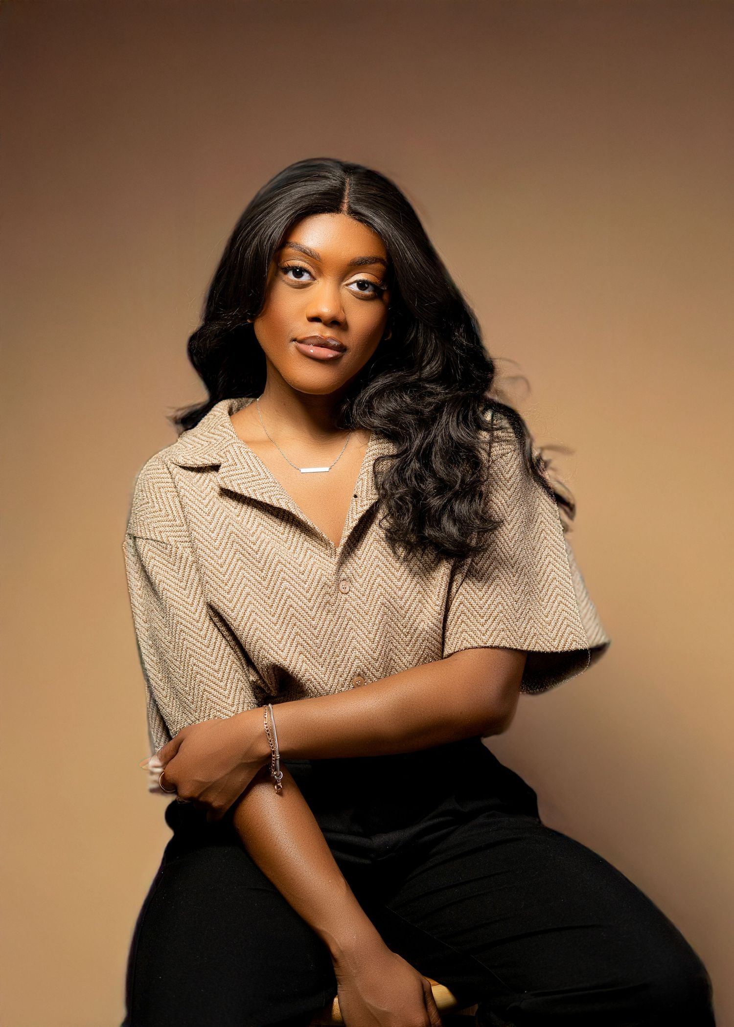 A young Black woman in a beige herringbone top and black pants poses confidently in a professional studio portrait.