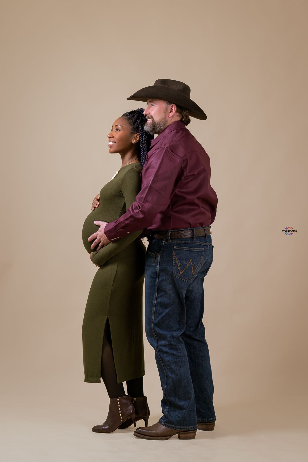 Pregnant interracial couple posing in studio, woman in olive dress, man in cowboy hat and burgundy shirt, maternity photo.