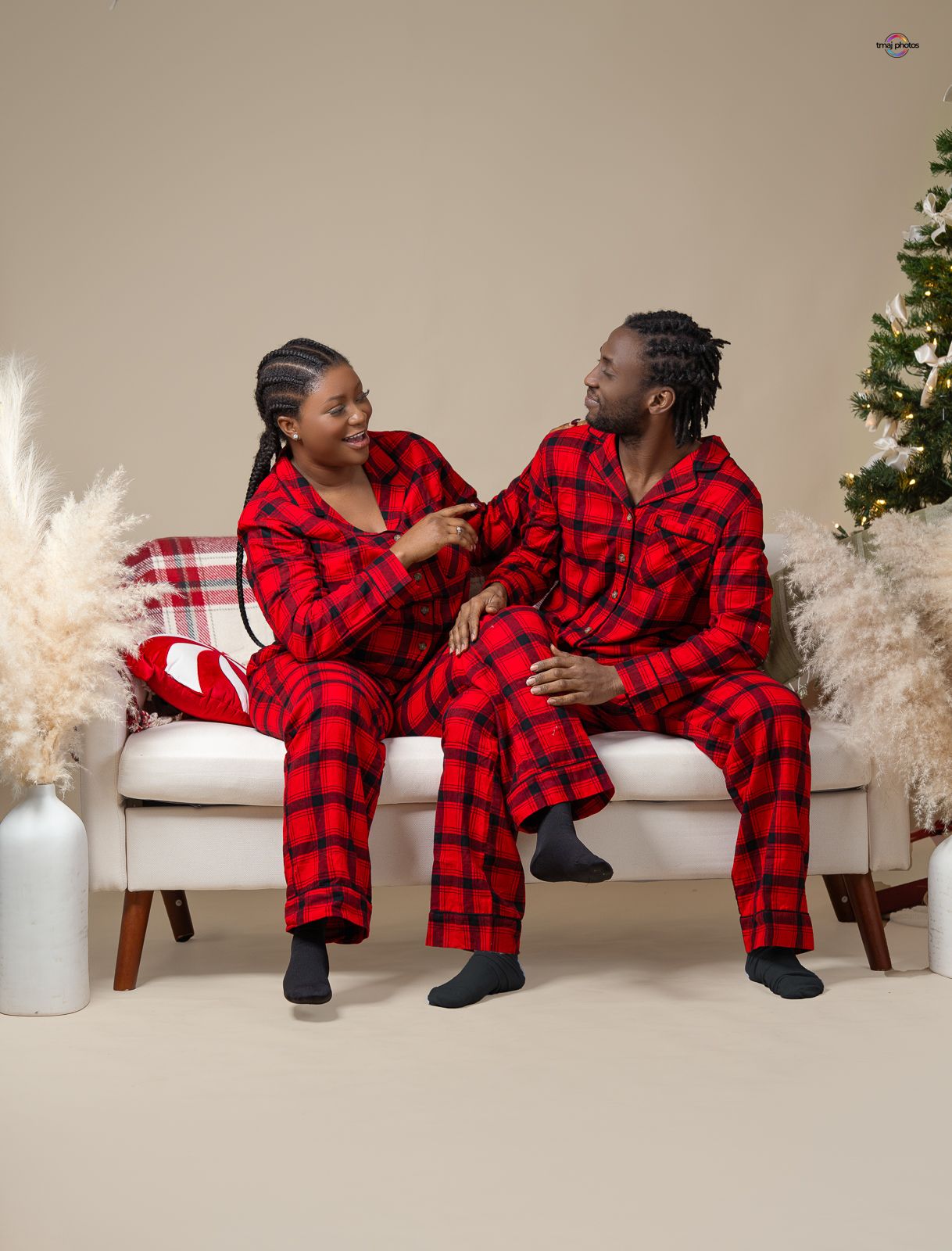 Couple in matching red plaid pajamas sitting on white sofa, smiling at each other near Christmas tree.