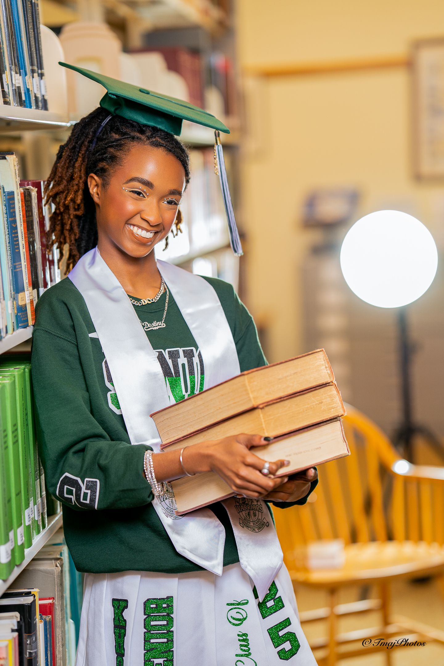 Smiling graduate in green cap and gown holding books in a library for senior portrait photos.