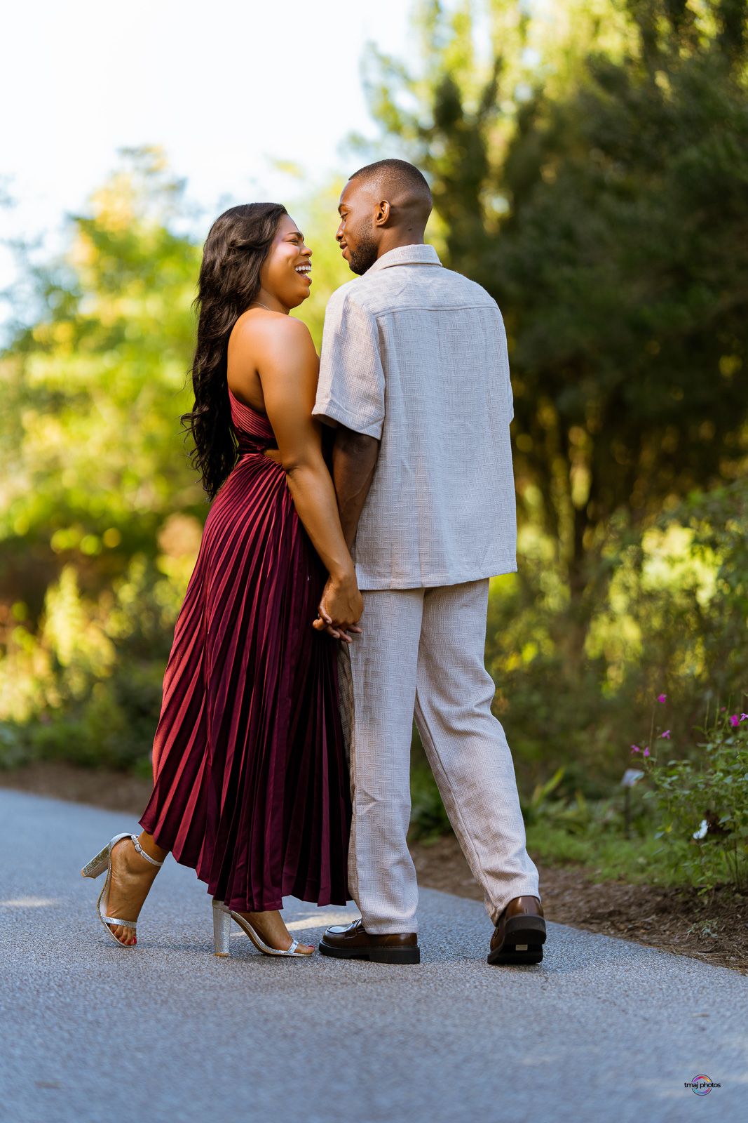 A couple holds hands and smiles at each other on a tree-lined path, dressed in elegant burgundy and linen outfits.