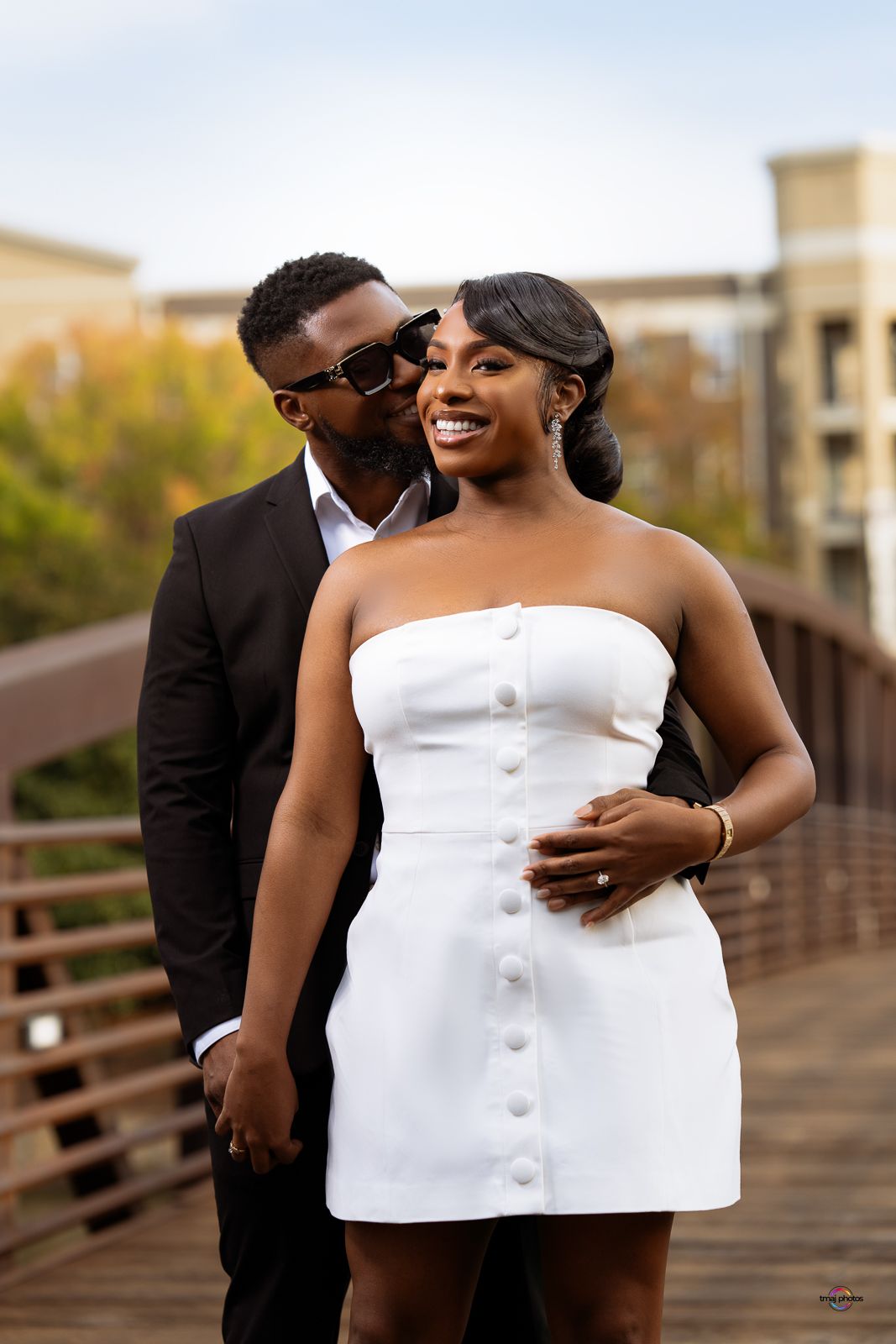A couple poses on a bridge, man in black suit kisses woman's cheek as she smiles in white mini dress.