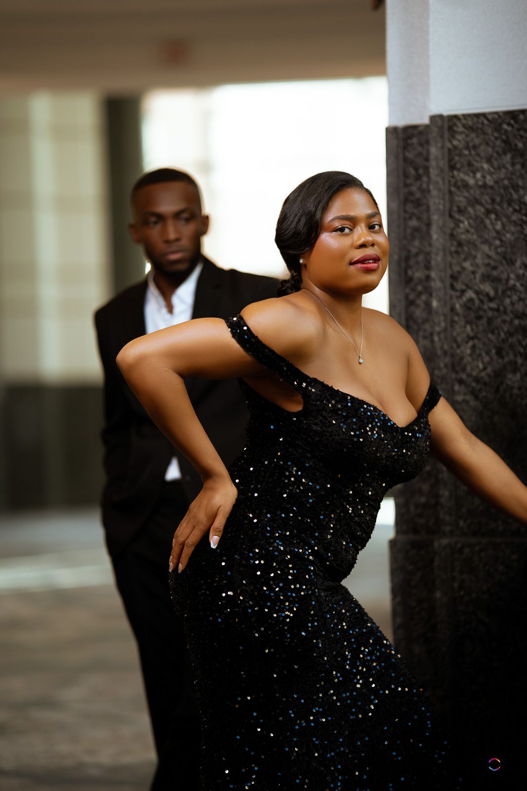A woman in a black sequin off-shoulder gown poses elegantly with a man in a suit standing behind her.