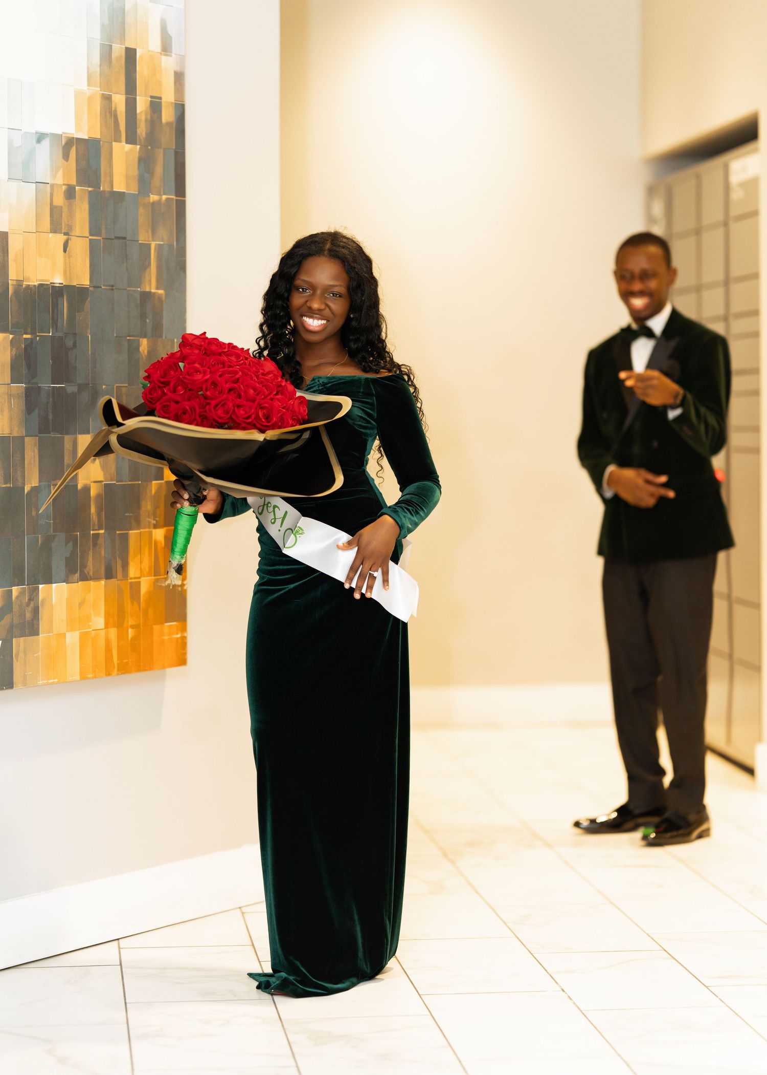 Woman in emerald green velvet gown smiles holding large heart-shaped red rose bouquet during proposal moment.
