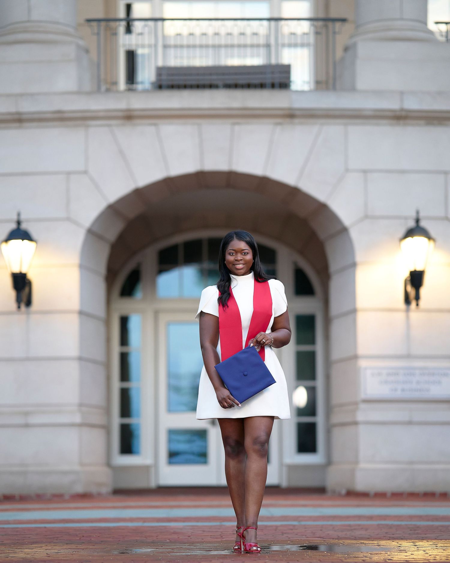 Young woman in white dress and red sash holds blue folder, standing before an arched building entrance.