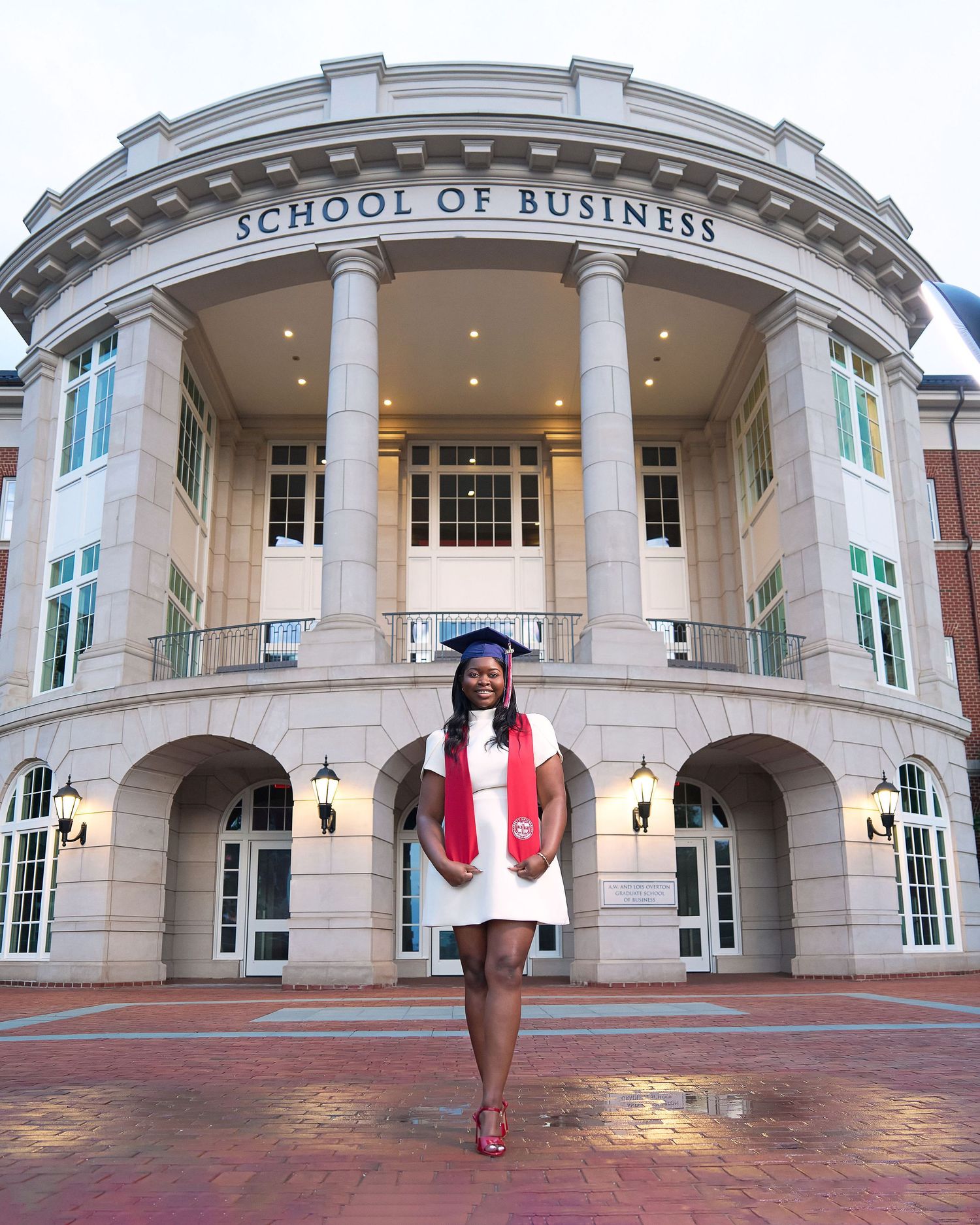 Graduate in cap and gown poses proudly in front of a School of Business building for graduation photos.