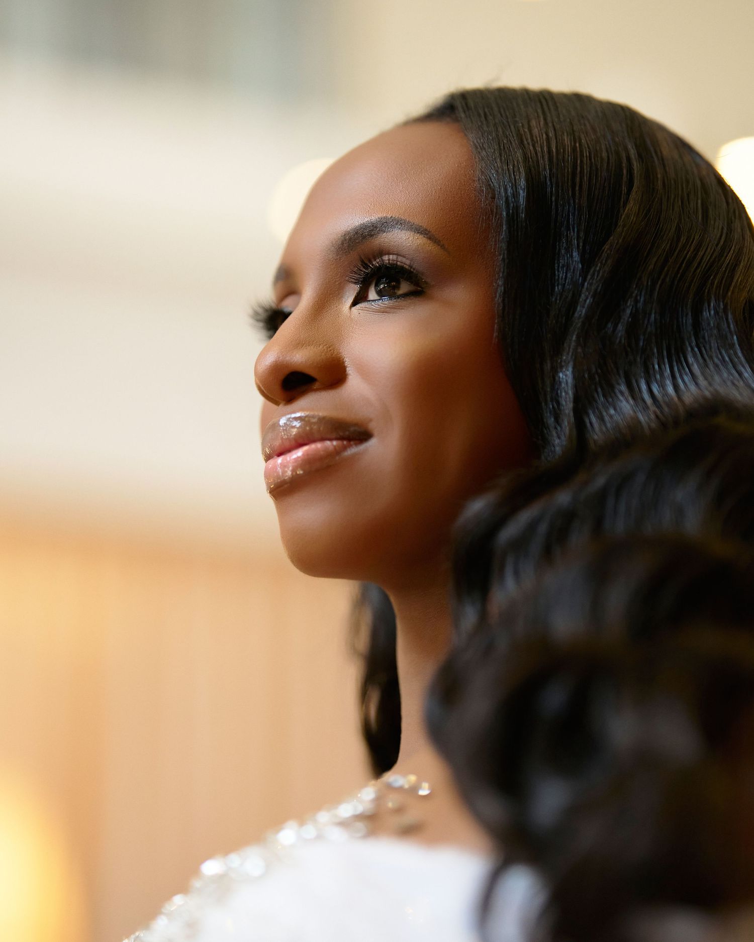 A Black woman with elegant wavy hair and glam makeup gazes upward, wearing a white embellished outfit at a formal event.