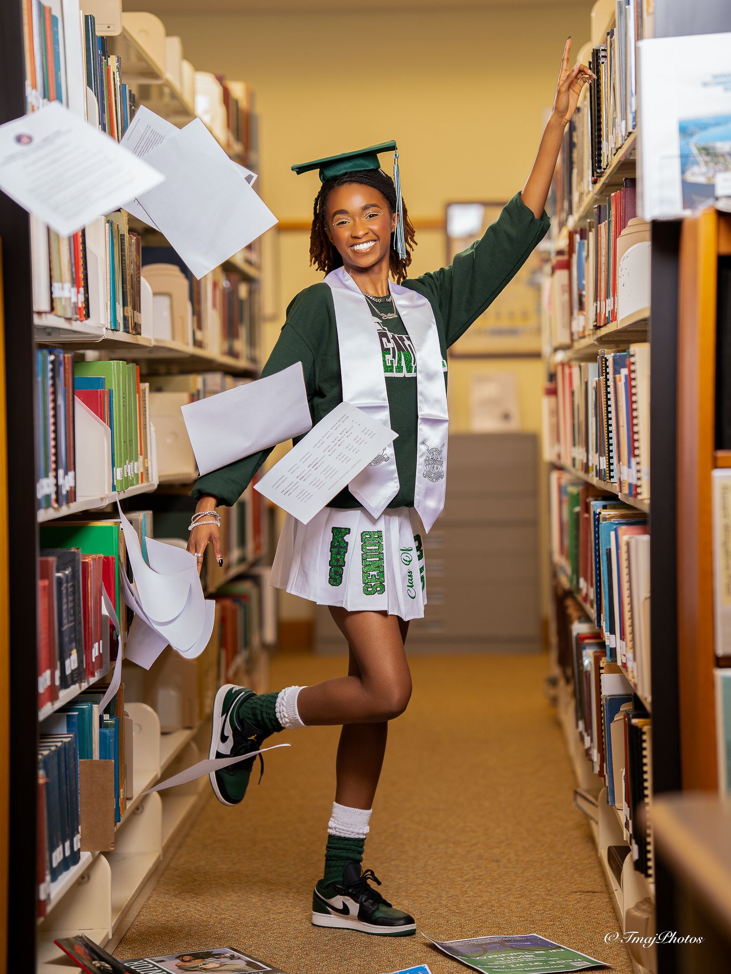 Joyful graduate tossing papers in library aisle wearing green cap and gown.