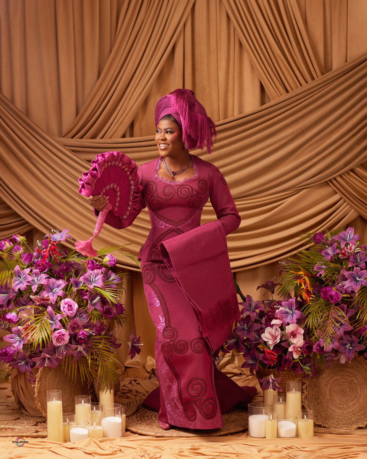 Woman in elegant magenta Nigerian attire with gele headwrap holds a decorative fan, surrounded by purple floral arrangements.